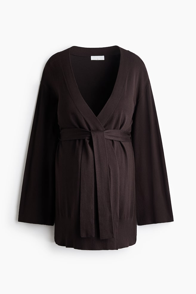 Un cardigan en maille marron foncÃ© est prÃ©sentÃ© de face, avec un col en V profond et une ouverture sur le devant fermÃ©e par une ceinture Ã  nouer assortie Ã  la taille. Le modÃ¨le met en valeur des Ã©paules tombantes, de longues manches larges avec poignets cÃ´telÃ©s, ainsi quâun bas de vÃªtement cÃ´telÃ©. - 1