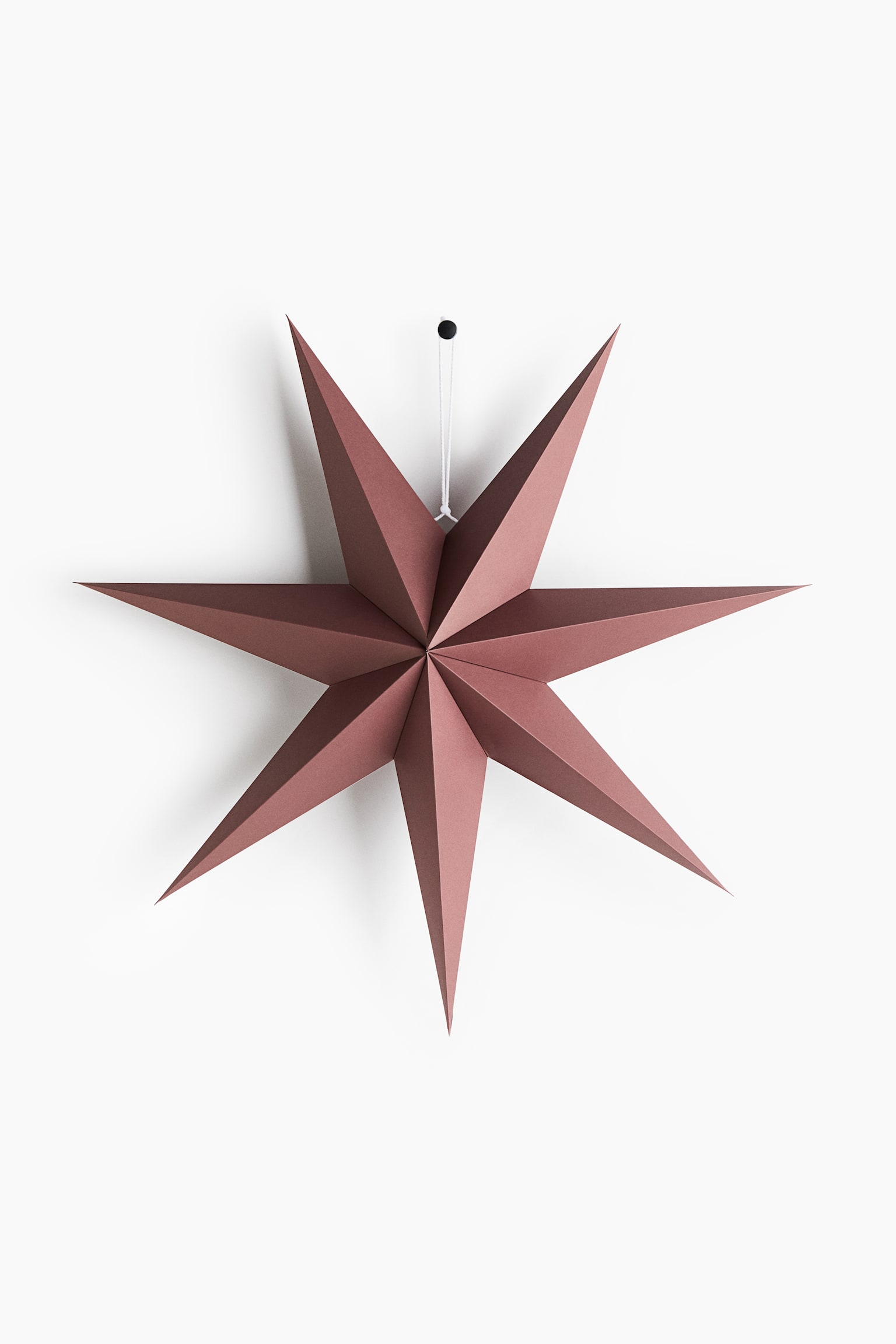 Uma decoraÃ§Ã£o em forma de estrela de papel, de cor rosa-pÃ³, estÃ¡ voltada para a frente, exibindo um design tridimensional de oito pontas com dobras geomÃ©tricas bem definidas. A estrela estÃ¡ pendurada por um fio fino branco preso a uma pequena mola preta. - 1