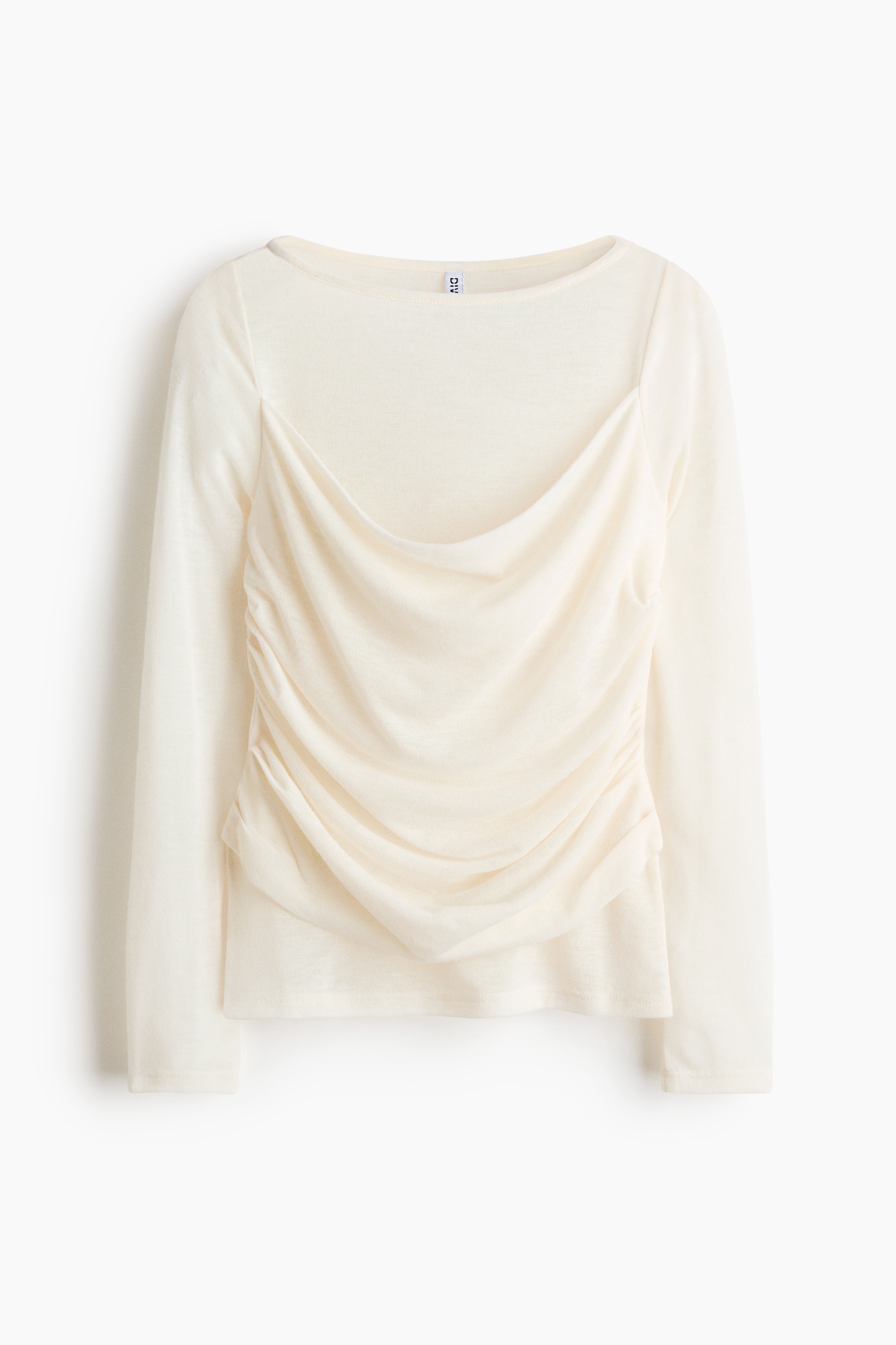 A cream-coloured, long-sleeved jersey top featuring a sheer boat neck and a draped, cowl-neck front layer is being worn by the model. The top is styled with light-wash, distressed denim jeans, with the model placing their hands in the front pockets.