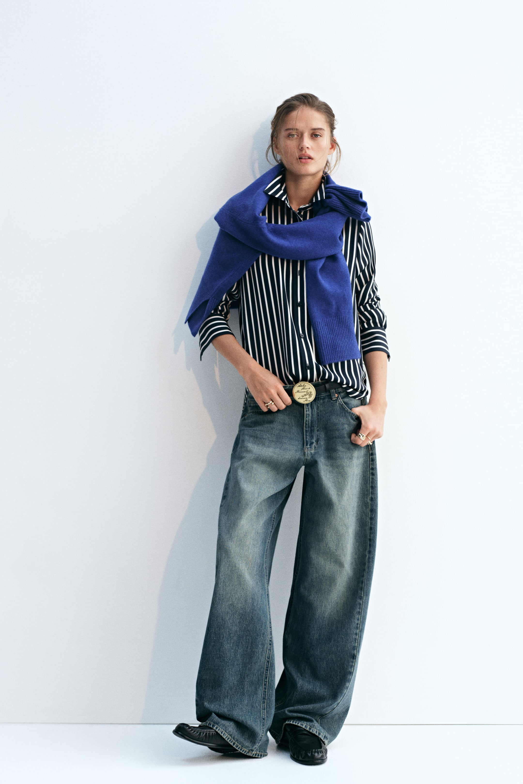 Ampliar la imagen: La modelo lleva una camisa con cuello a rayas en un patrón vertical en blanco y negro, parcialmente metida dentro de unos jeans de pierna ancha. La camisa de manga larga con botones está estilizada con un suéter tejido de color azul brillante colocado sobre los hombros.