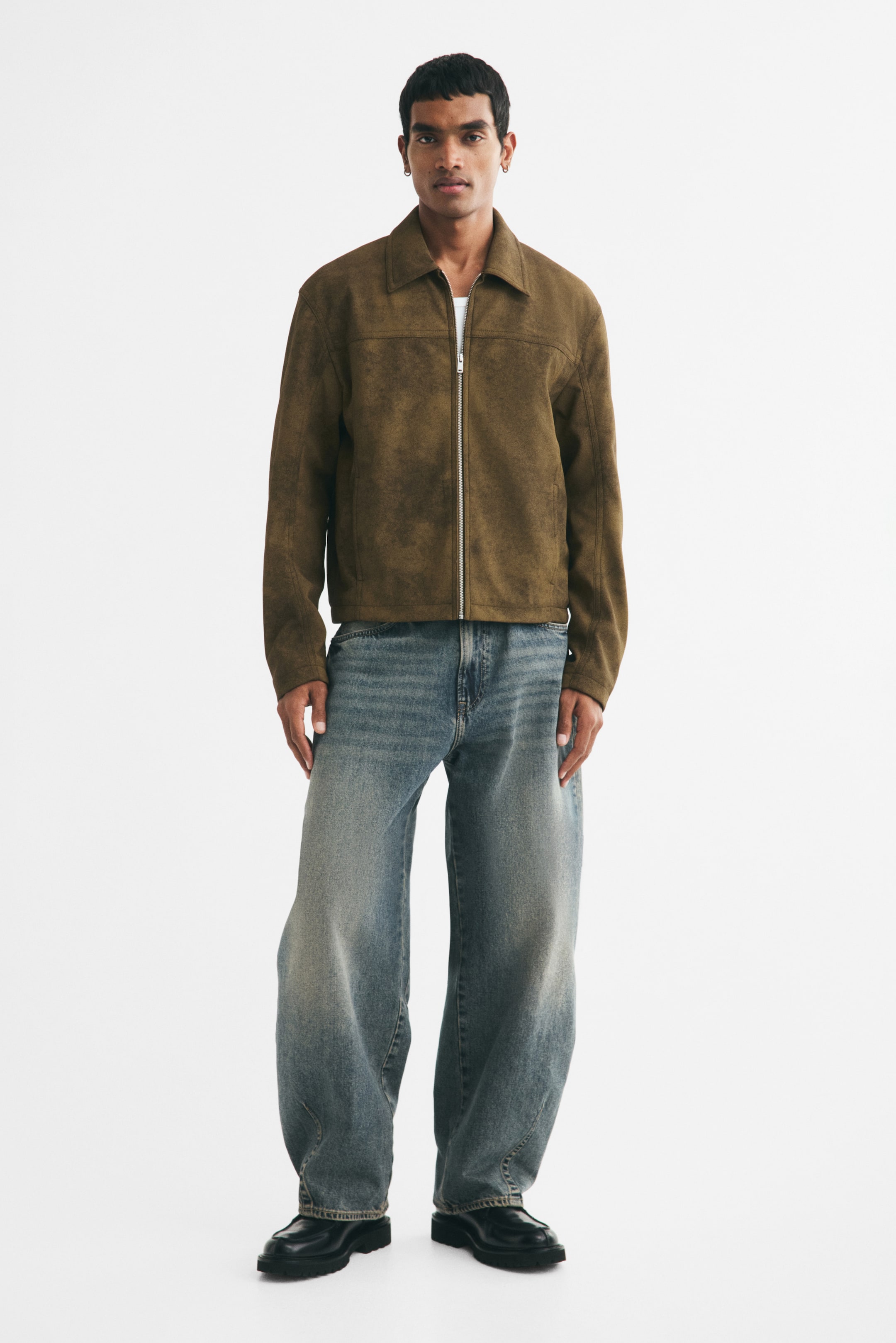View larger image: The model is wearing a brown suede-textured jacket with a classic collar, silver-toned front zip closure, and long sleeves. It is layered over a white T-shirt and paired with loose-fitting blue jeans.