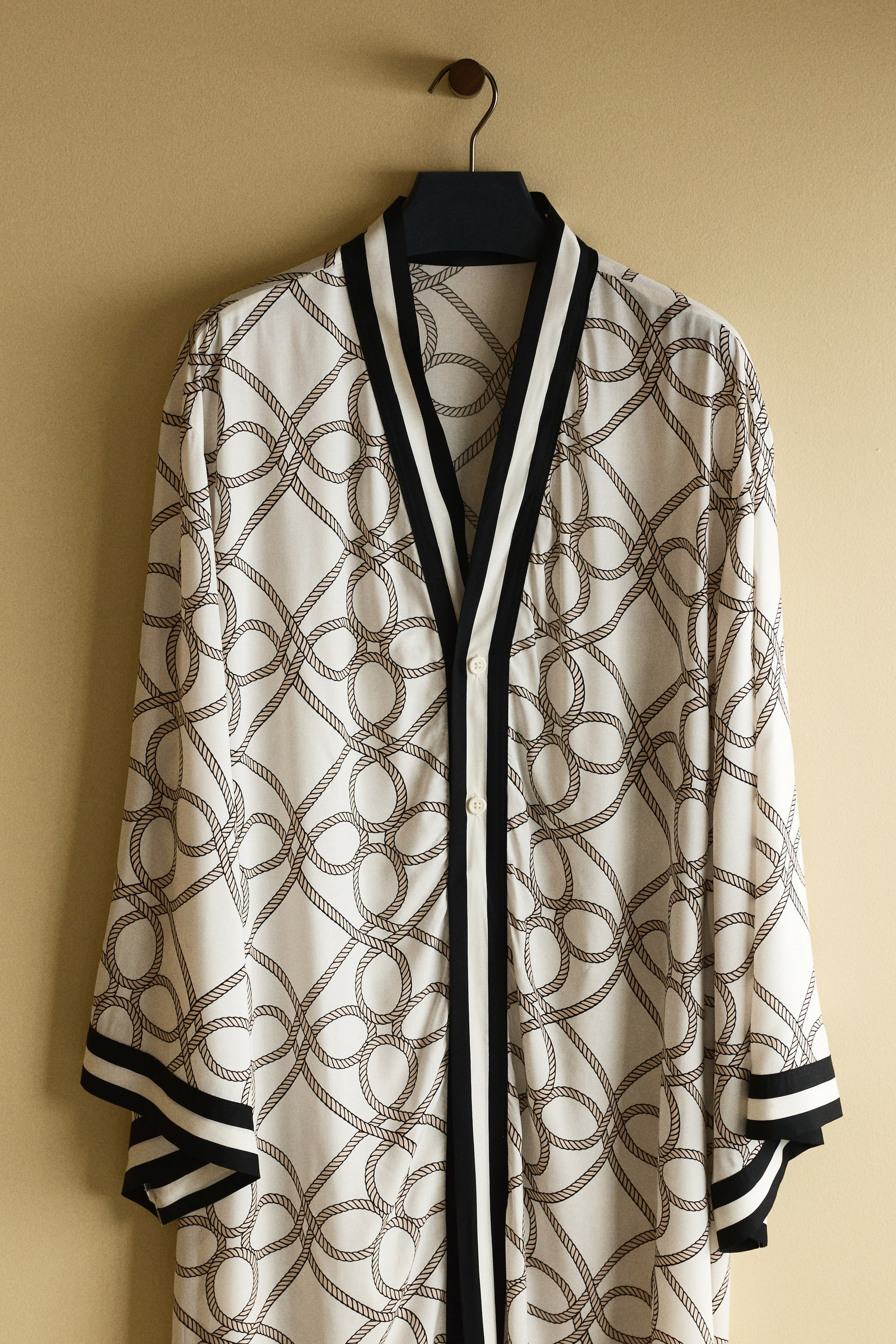 View larger image: A long white garment, showcasing an intricate brown rope pattern, is hanging facing forward. It features a deep V-neckline and front placket with wide black and white striped lapels, complemented by two visible white buttons, and its long sleeves are adorned with matching black and white striped bands.