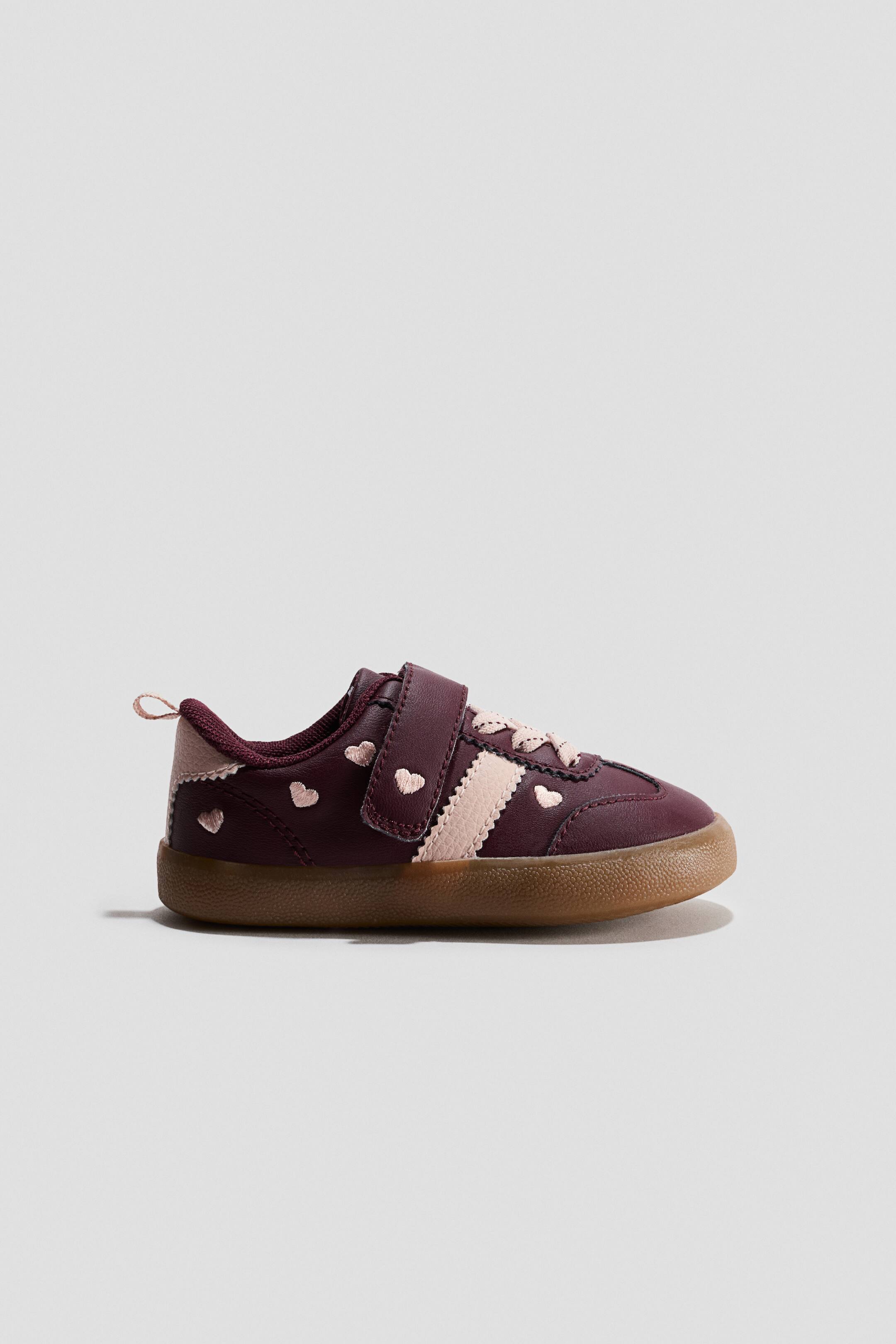 View larger image: Barefoot trainers in burgundy leather are shown in side profile, featuring a thin, flexible gum sole and a single hook-and-loop strap closure. The shoe displays decorative details, including small embroidered pink hearts, a textured pink side stripe with scalloped edges, and light pink laces.
