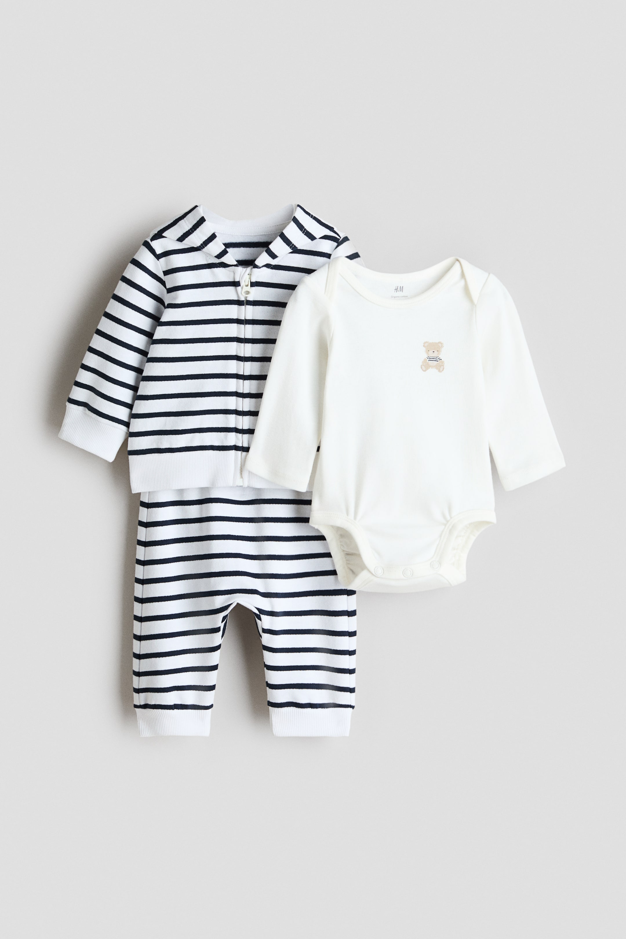 View larger image: A three-piece baby clothing set featuring a white and navy striped zip-through hoodie and matching joggers, presented alongside an off-white long-sleeved bodysuit. The striped hoodie, shown front-on, displays a front zip with ribbed cuffs and hem, while the matching joggers show a ribbed waist and ribbed hems. The off-white bodysuit, also shown front-on, presents lapped shoulders and a small teddy bear print on its chest.