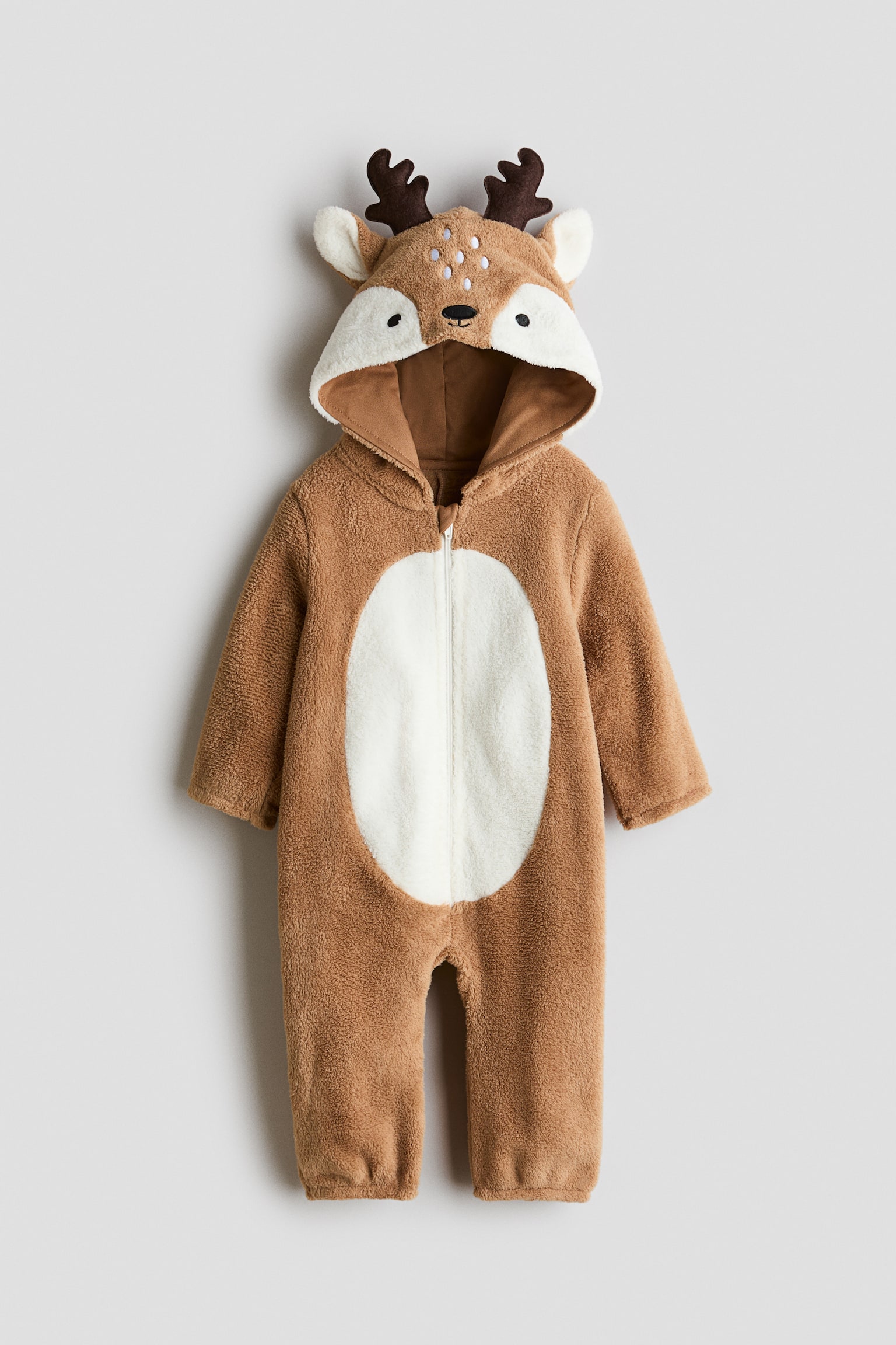 This fancy dress costume, designed as a plush brown deer onesie, is facing front and displaying a full-length front zipper with a contrasting white oval belly patch. The attached hood is showcasing an embroidered deer face with white spots, fabric ears, and dark brown antlers, while the long sleeves and legs are ending in elasticized cuffs.
