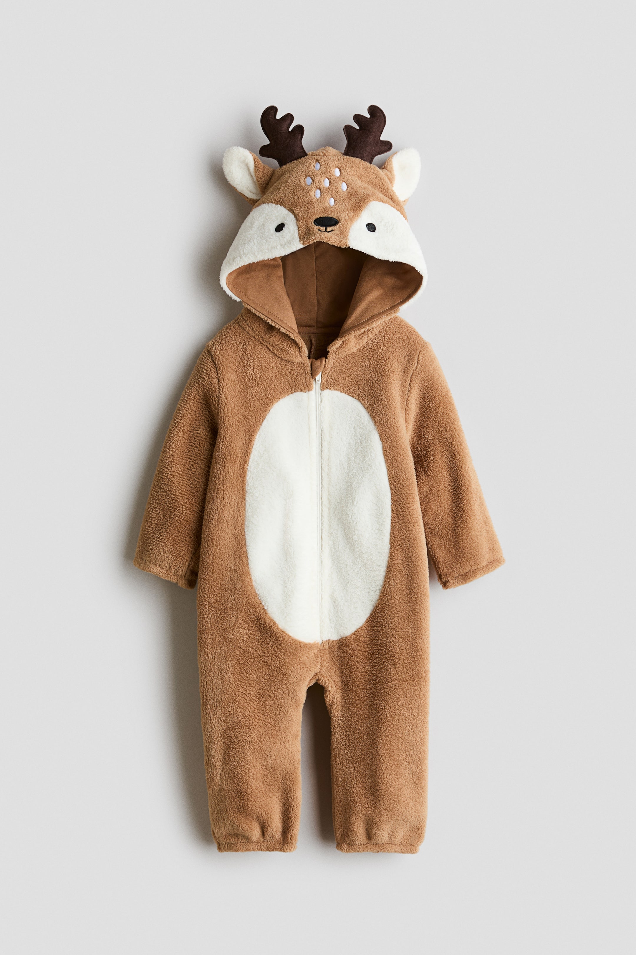 Voir une image plus grande: Ce costume déguisé raffiné, conçu comme une combinaison une pièce en peluche brun cerf, est présenté de face et met en valeur une fermeture éclair frontale sur toute la longueur avec un appliqué ovale blanc contrastant au niveau du ventre. La capuche intégrée arbore un visage de cerf brodé avec des taches blanches, des oreilles en tissu et des bois brun foncé, tandis que les longues manches et les jambes se terminent par des poignets élastiqués.