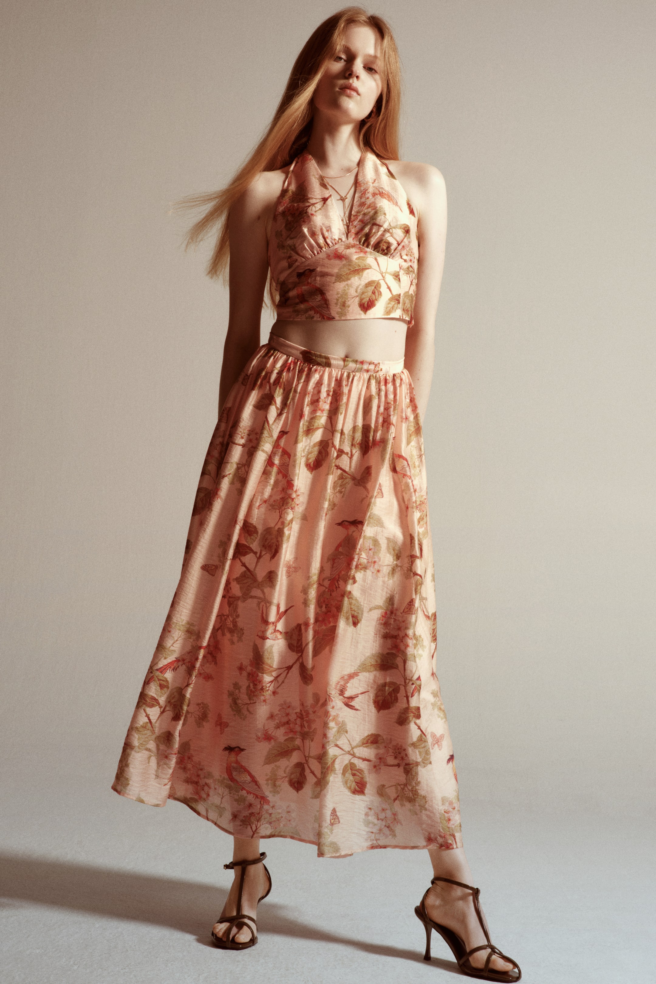 View larger image: The model is wearing an ankle-length, flared skirt in a light pink printed weave. The skirt boasts a high waist with a narrow gathered waistband, adorned with a floral and bird motif, and is styled with a matching cropped halter top.