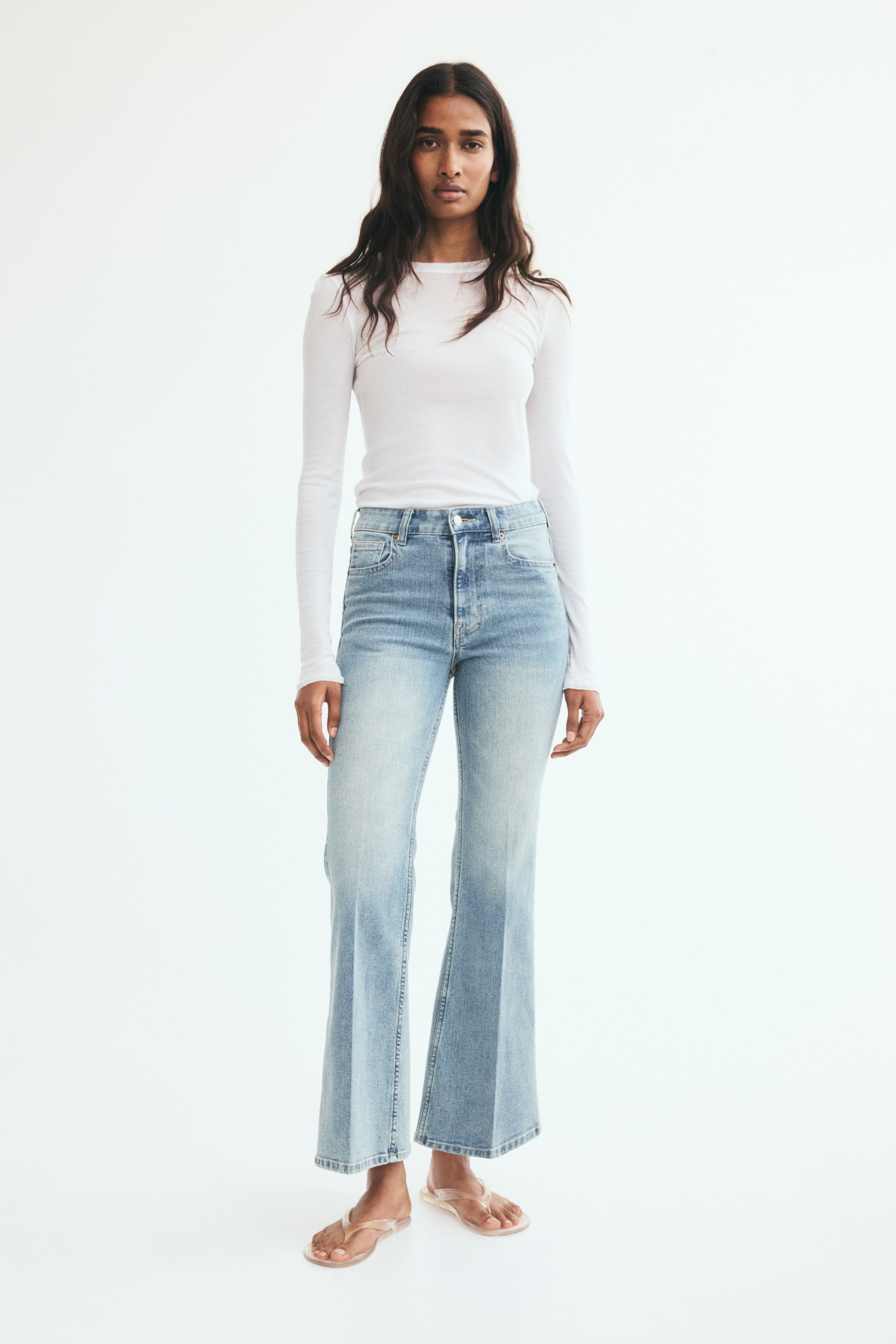View larger image: The model is wearing light-wash blue denim jeans with a high waist, a slim fit through the thighs, and flared legs. The jeans feature vertical creases down the front of the legs and are styled with a tucked-in, long-sleeved white top.