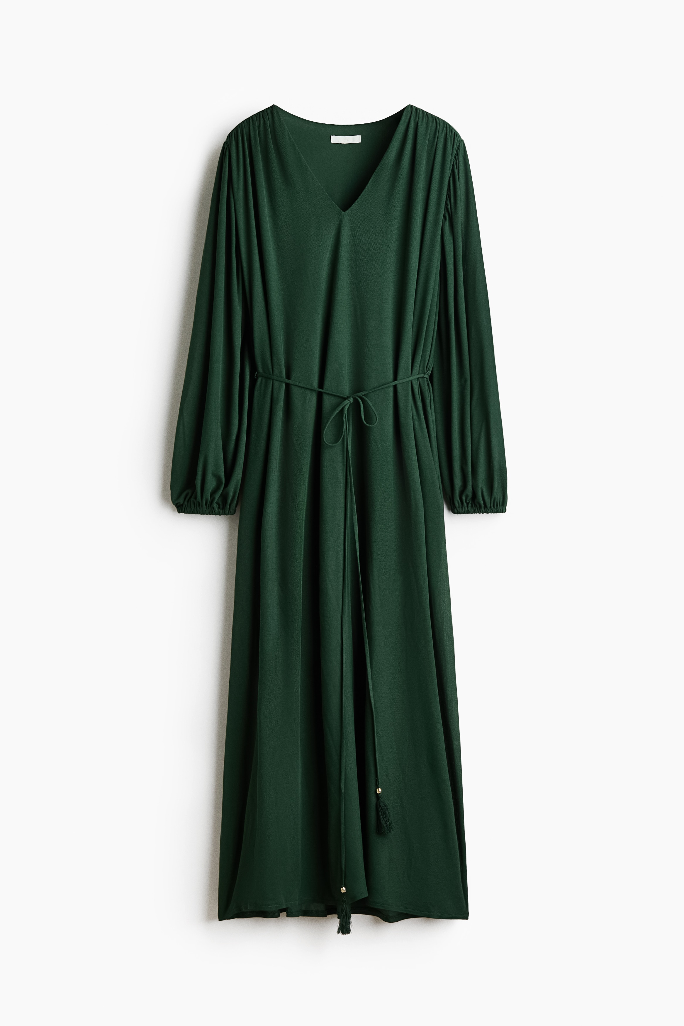 A long, loose-fit dark green dress in fine ribbed jersey is being worn by the model, who is standing in profile with one hand resting on her hip. The dress features long balloon sleeves with elasticated cuffs and a thin tie belt at the waist with tasselled, beaded ends.