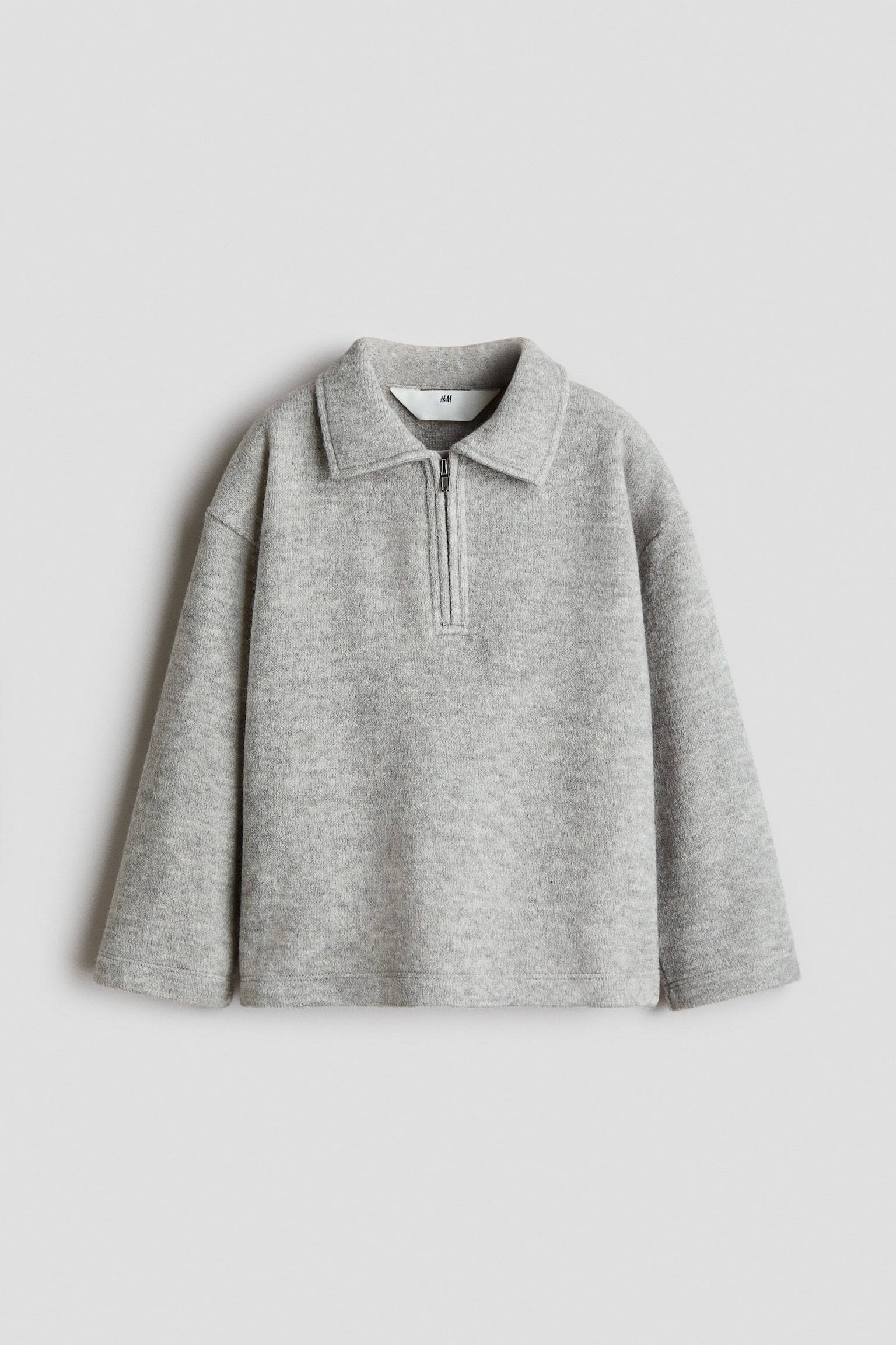 A light grey half-zip pullover is facing front, showcasing a soft, textured fleece-like material and a relaxed silhouette. It features a pointed collar, a short silver-tone zip, dropped shoulders, and wide, straight-cut long sleeves.