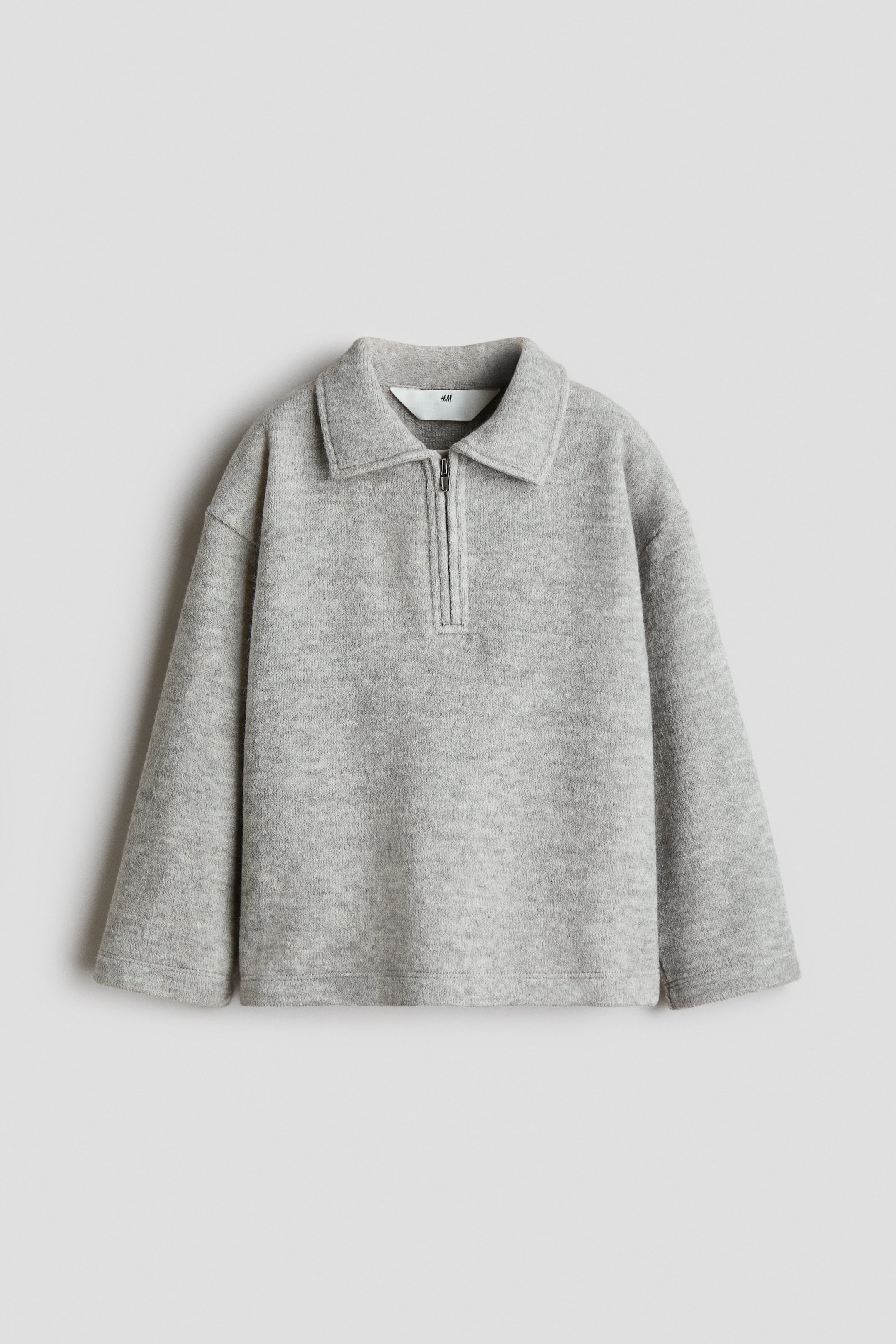 Grotere afbeelding bekijken: Een lichtgrijze pullover met halve rits is van voren te zien, met een zachte, gestructureerde fleece-achtige stof en een ontspannen silhouet. Het model heeft een puntkraag, een korte rits in zilverkleur, verlaagde schouders en wijde, rechte lange mouwen.