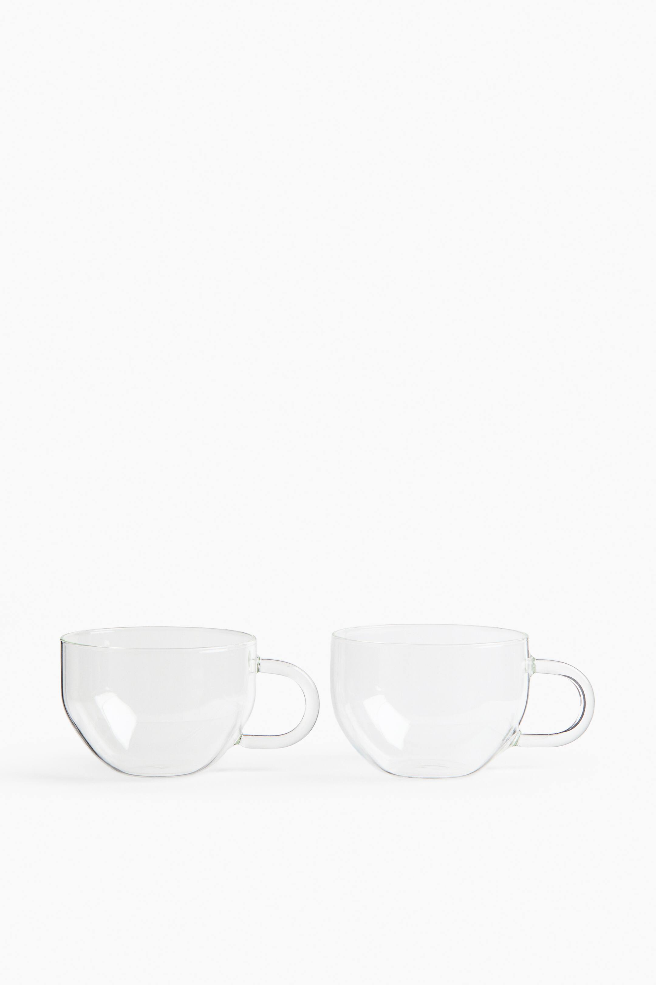 View larger image: Two clear glass cups are standing side-by-side, each featuring a smooth, round, bowl-like body and a small, C-shaped handle.