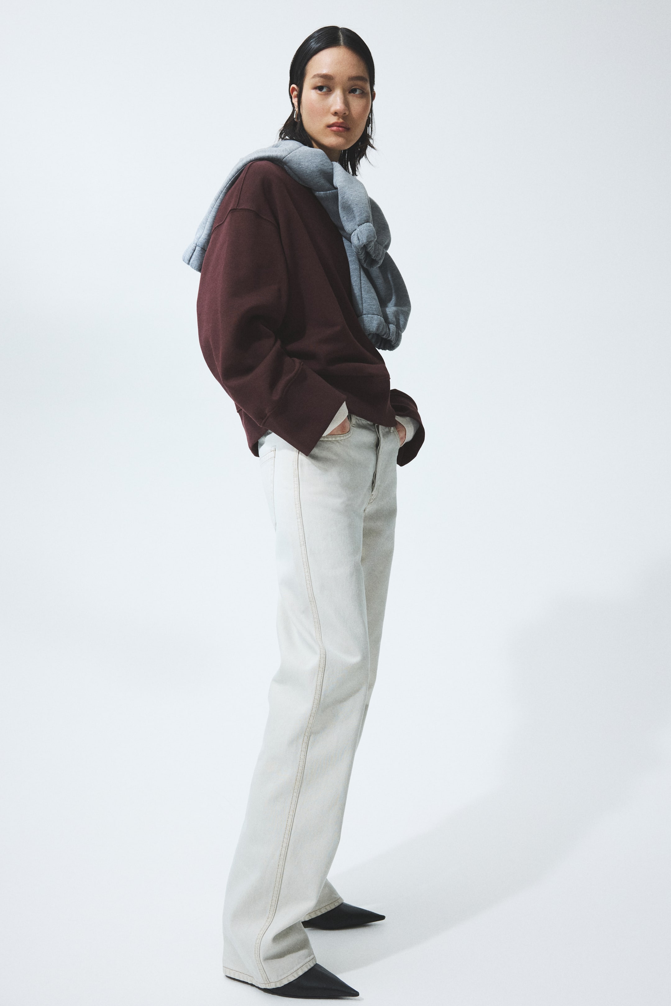 הצגת תמונה גדולה יותר: סווטשירט בורדו גדול במיוחד, עם חפתים רחבים ושוליים רפויים, נלבש מעל חולצת תחתית לבנה ומעוצב עם סווטשירט אפור בהיר שמונח על הכתפיים, כאשר השרוולים קשורים מלפנים. מכנסי ג'ינס בגוון בז' בהיר בגזרה ישרה עם תפרים אנכיים נראים לעין נלבשים עם נעליים שחורות עם חוד מחודד.