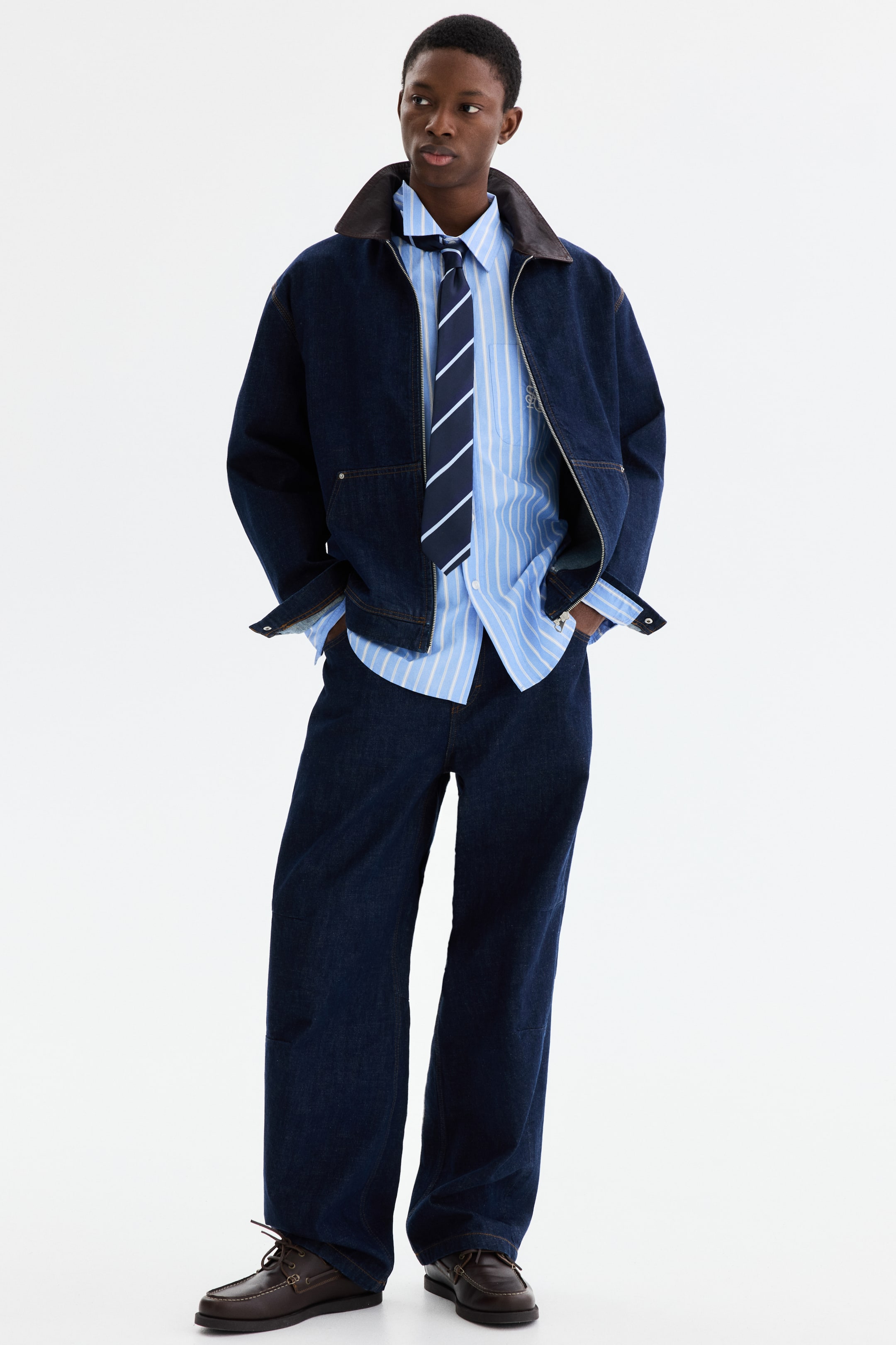 Agrandir l'image: Une veste en denim bleu foncé avec un col à revers marron et des fermetures éclair argentées est superposée à une chemise à rayures bleu clair et blanches et une cravate à rayures bleu marine et bleu clair. Cet ensemble est assorti d'un pantalon en denim bleu foncé à jambes larges et de chaussures bateau en cuir marron foncé avec des coutures apparentes.