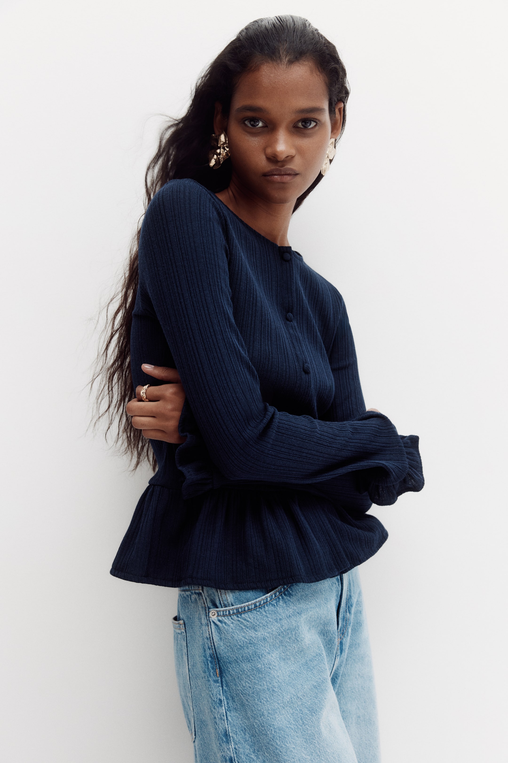 View larger image: A navy-blue ribbed-knit top features a round neck, button-front closure and long sleeves with ruffled cuffs. The fitted bodice flares into a ruffled peplum hem, sitting just above light-blue jeans.
