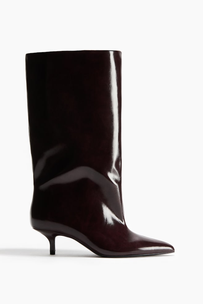 Ein dunkel bordeauxfarbener Stiefel aus glänzend beschichtetem Stoff ist in seitlicher Profilansicht nach rechts ausgerichtet zu sehen und verfügt über eine spitze Zehenpartie, einen niedrigen, schmalen Absatz und einen weiten, knöchelhohen Schaft. Die stark reflektierende Oberfläche betont die schlanke Struktur des Stiefels sowie die leichte Fältelung am Knöchel. - 1