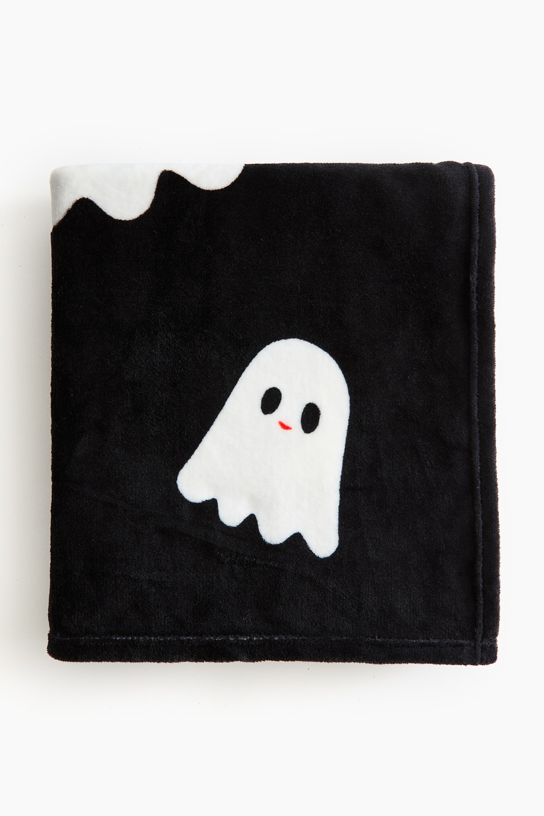 View larger image: A black fleece blanket is lying flat, featuring a white ghost appliqué with black oval eyes and a red curved mouth. A wavy white pattern is also visible along the blanket's top left edge.