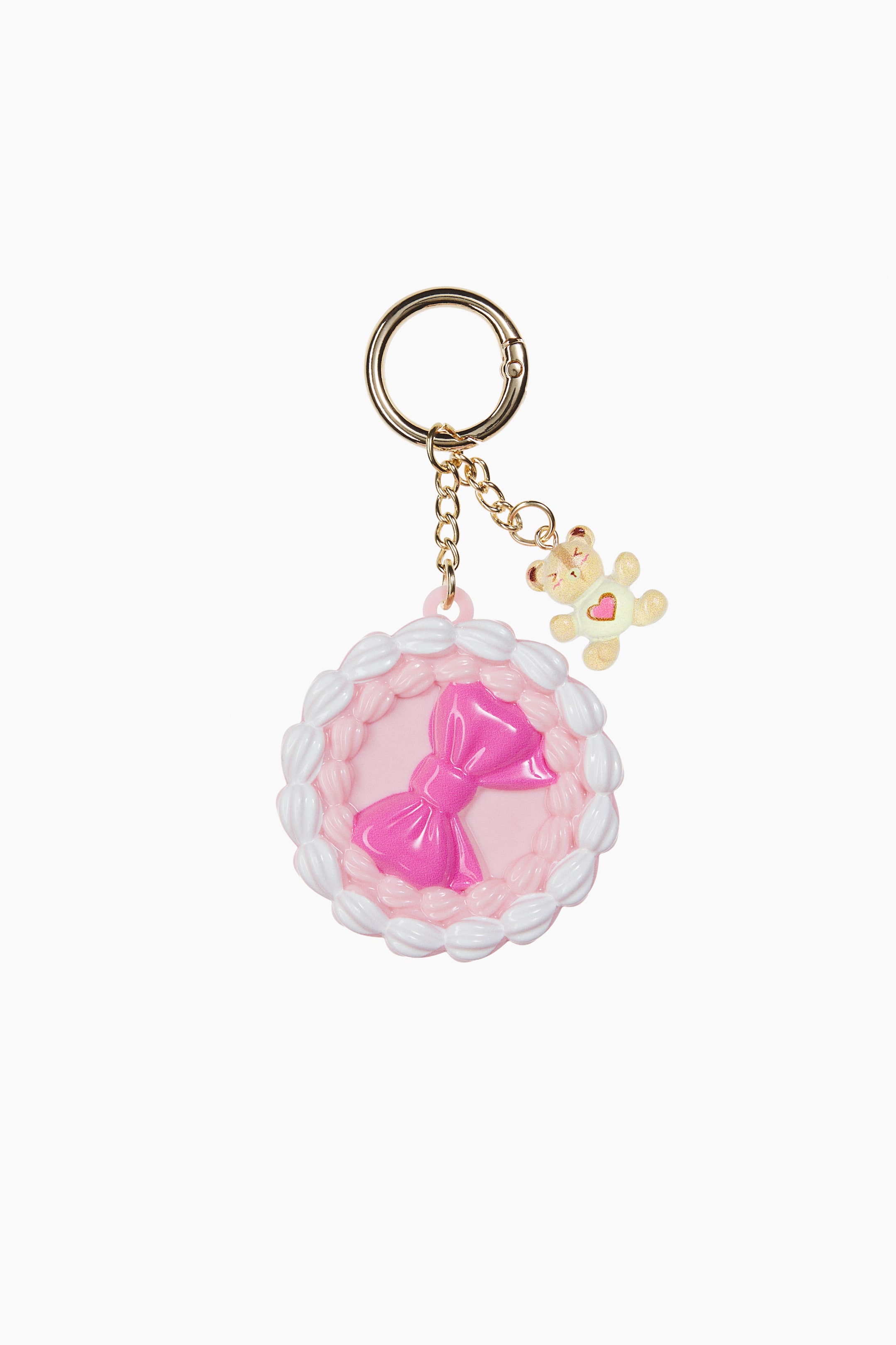 View larger image: A round, cake-shaped lip and cheek balm container is shown from the front, featuring a central pink bow and a white frosted-style border. The container hangs from a gold-toned round clasp and chain, which also holds a small teddy bear charm decorated with a pink heart.