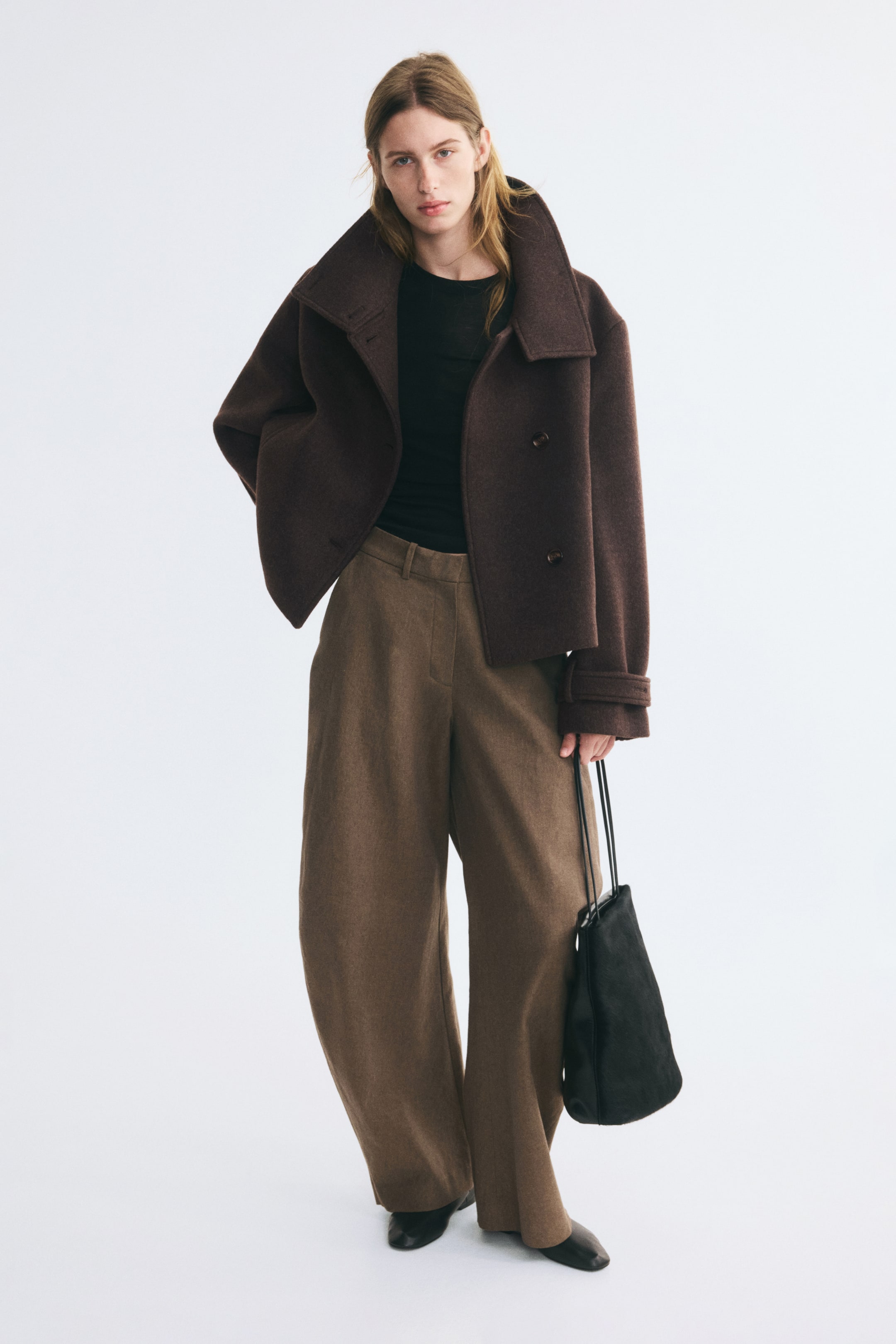 Visa större bild: En oversized, mörkbrun jacka i ett filtad ullblandat tyg bärs öppen av modellen. Jackan har en bred, strukturerad krage, knäppning framtill med knappar och ärmslut med knappflikar, stylad över en svart topp och bruna vida byxor.
