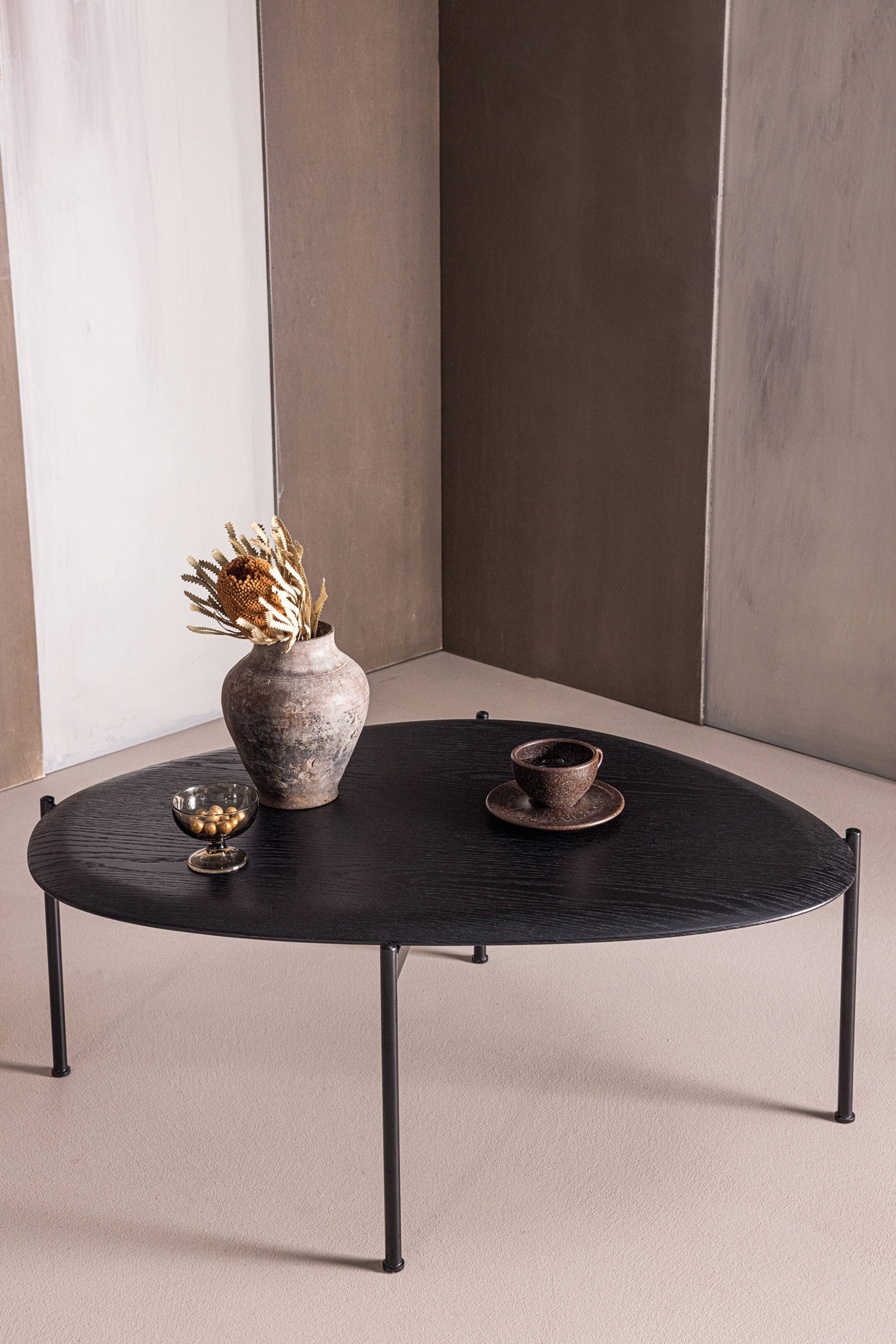 Grotere afbeelding bekijken: Een zwarte onregelmatige ovale salontafel met een houten blad met zichtbare nerf, ondersteund door vier dunne, gespreide zwarte metalen poten. Op het oppervlak staat een rustieke vaas met gedroogde botanicals, een klein glas en een getextureerde donkere kop met bijpassend schoteltje.
