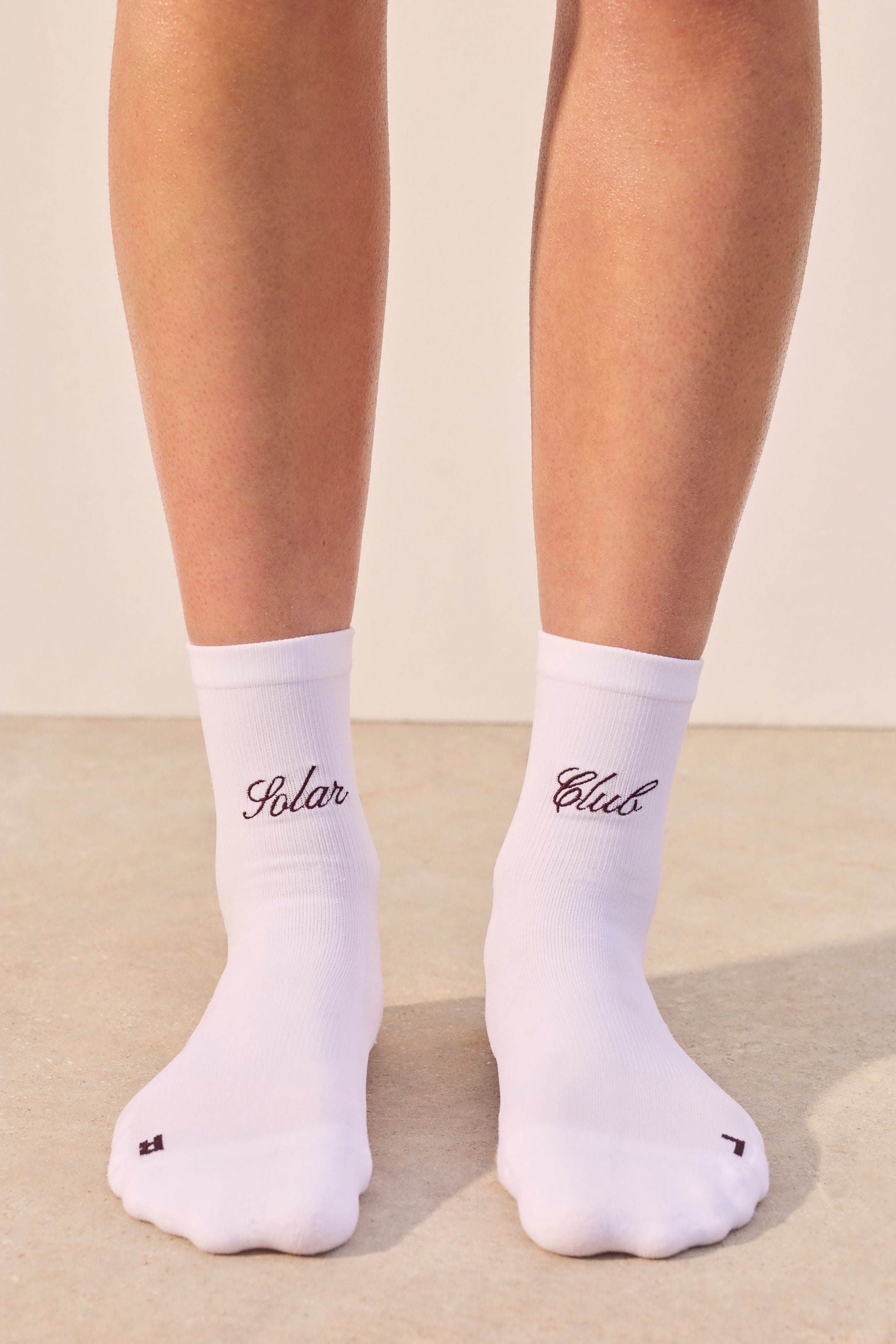 View larger image: A pair of white socks is worn, facing forward, with each sock showcasing a ribbed cuff. The left sock features "Solar" embroidered in black script on its outer ankle and a small black square graphic on its toe, while the right sock features "Club" embroidered in black script on its outer ankle and a small black L-shaped graphic on its toe.
