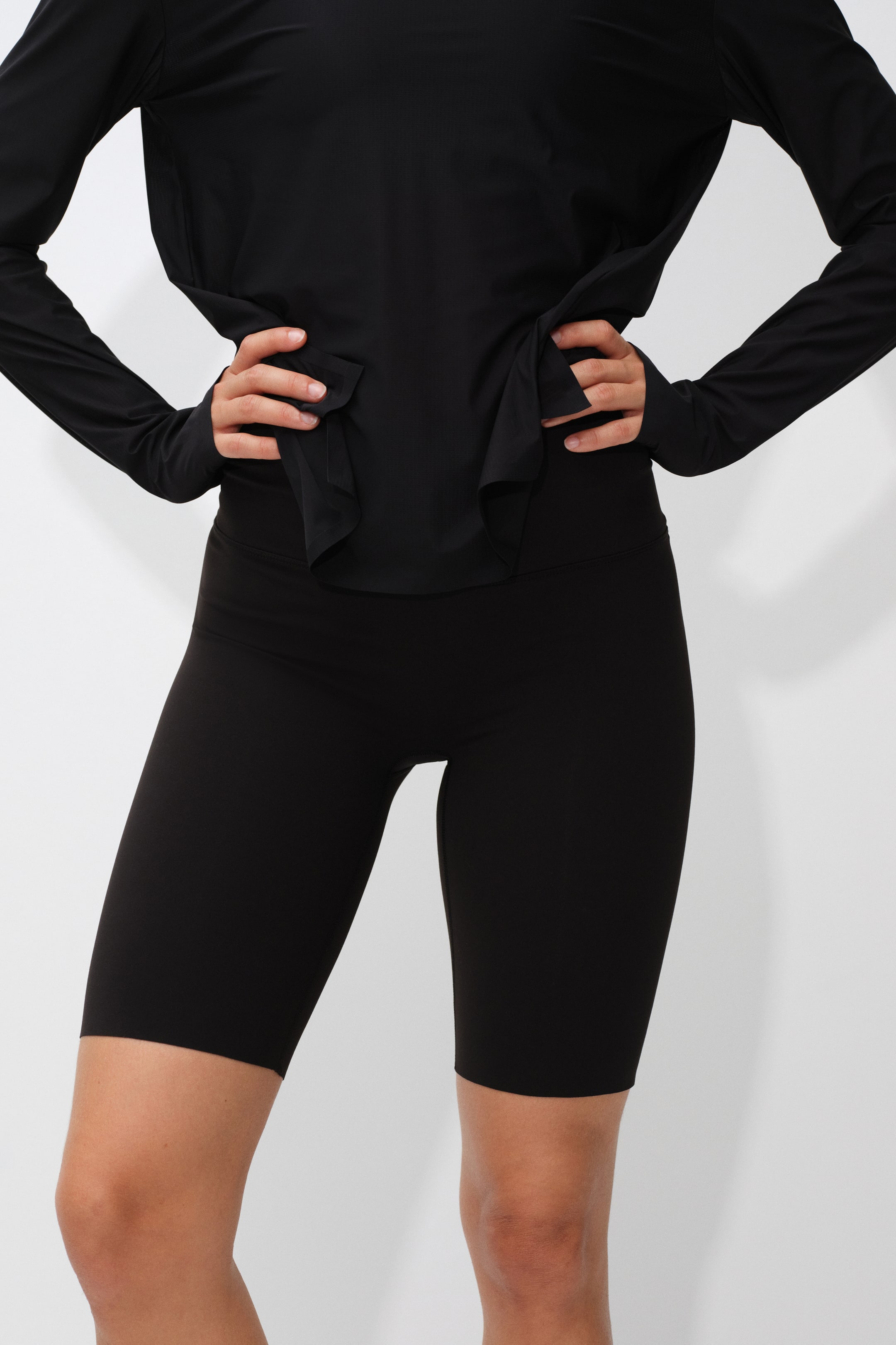 View larger image: The model is wearing black high-waisted bike shorts paired with a matching black long-sleeve athletic top. She is posing with her hands on her hips, highlighting the close fit and mid-thigh length of the shorts.