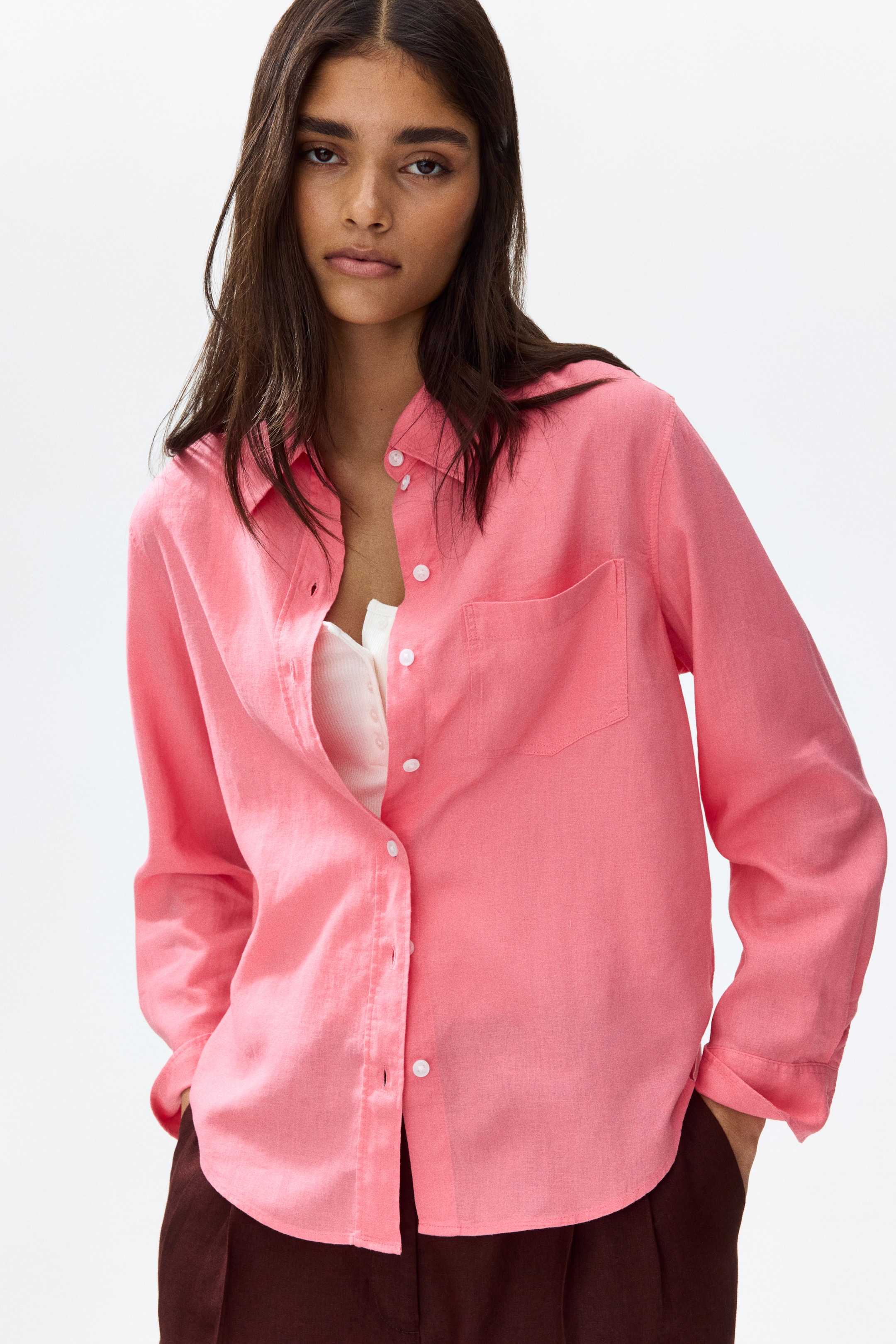 View larger image: The model is wearing a pink loose-fit shirt crafted from an airy weave, featuring a collar and white buttons running down the front. It includes an open chest pocket. The top buttons are undone, revealing a white ribbed top underneath. The long sleeves are casually pushed up, showcasing the shirt's curved hem.