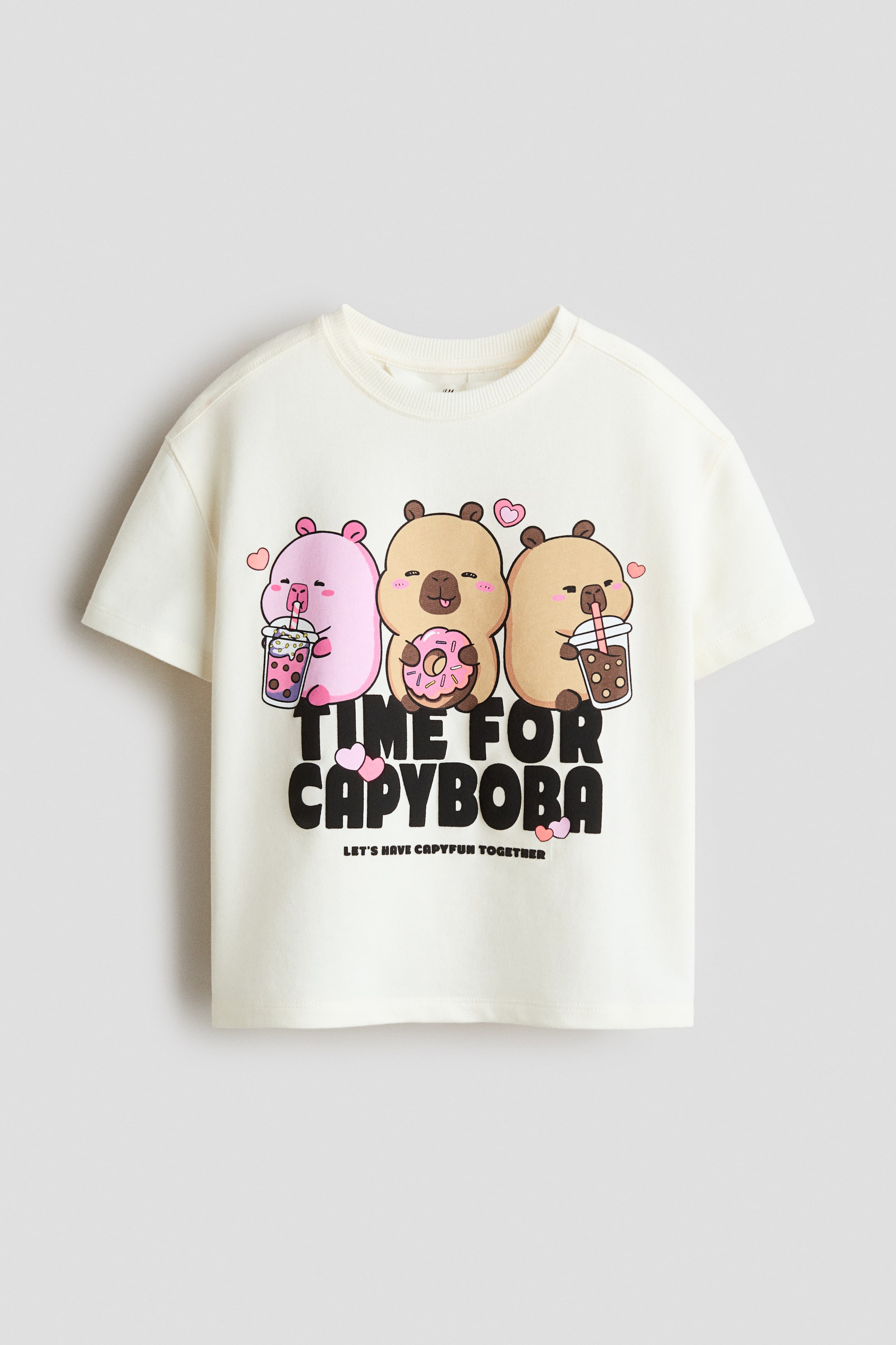 Grotere afbeelding bekijken: De voorkant van de crèmekleurige top met korte mouwen, gemaakt van sweatshirtstof, heeft een ronde, geribbelde halslijn en verlaagde schouders. Het toont een grafische print van drie cartoon capibara's die snacks en drankjes vasthouden, met tekst die luidt "TIME FOR CAPYBOBA" en "LET'S HAVE CAPYFUN TOGETHER," geaccentueerd door roze hartjes.