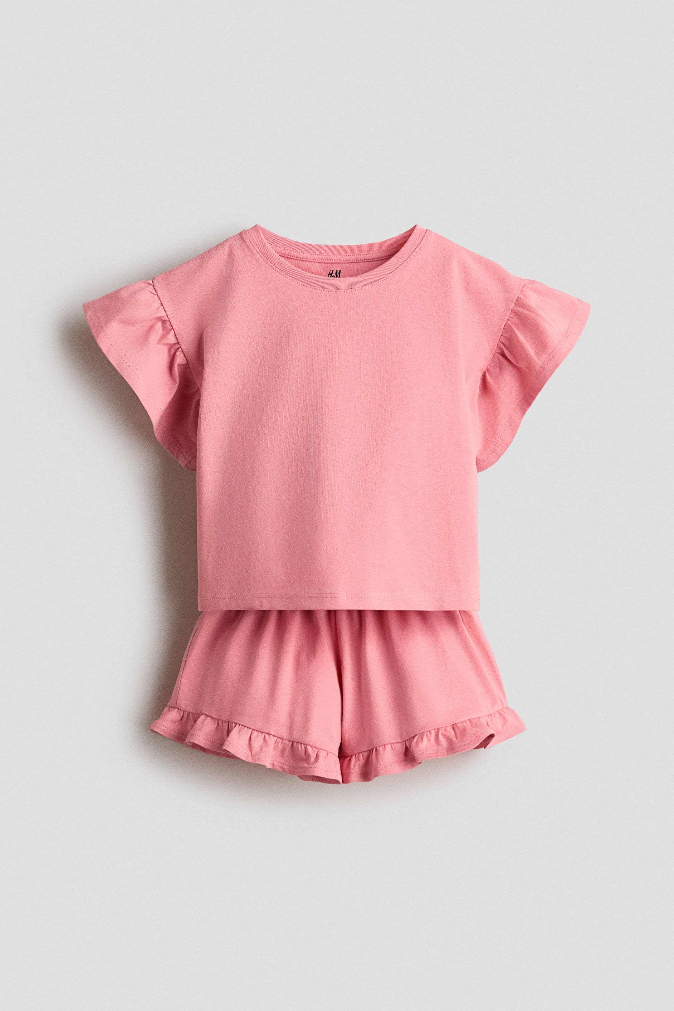 View larger image: A pink cotton jersey set showcases a top and shorts, viewed from the front. The top features a round neckline, dropped shoulders, and flutter sleeves, while the shorts are designed with an elastic waistband and frill-trimmed hems.