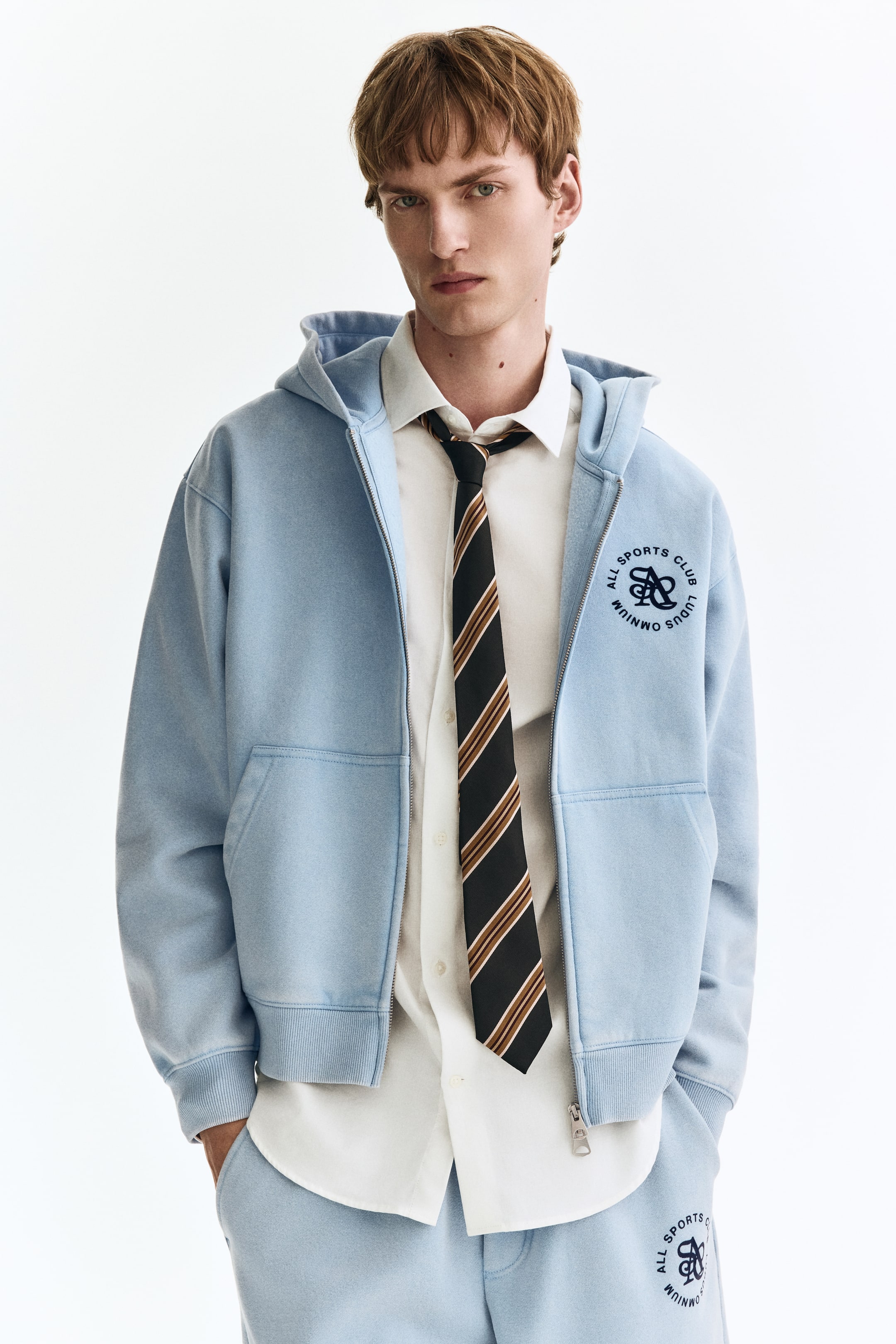 Visa större bild: En ljusblå zip-up hoodie bärs öppen, med en huva, långa ärmar med ribbade muddar, en ribbad fåll och en broderad cirkulär logotyp 'ALL SPORTS CLUB LUDUS OMNIUM' på vänster bröst. Denna hoodie är lagrad över en vit skjorta med krage och en svart och brunrandig slips, med matchande ljusblå träningsbyxor som också bärs, med en liknande broderad logotyp på vänster lår.