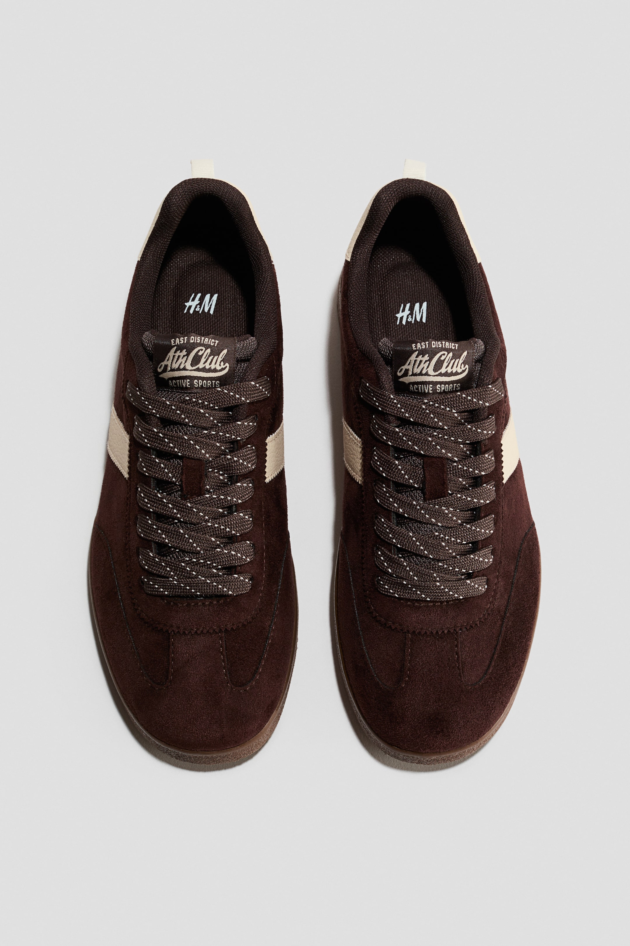 View larger image: A pair of dark brown suede sneakers is showcased from a top-down front view. Each sneaker features light beige stripes on its sides, dark brown speckled laces, and a tongue label displaying "H.M" and "Ath Club" branding.