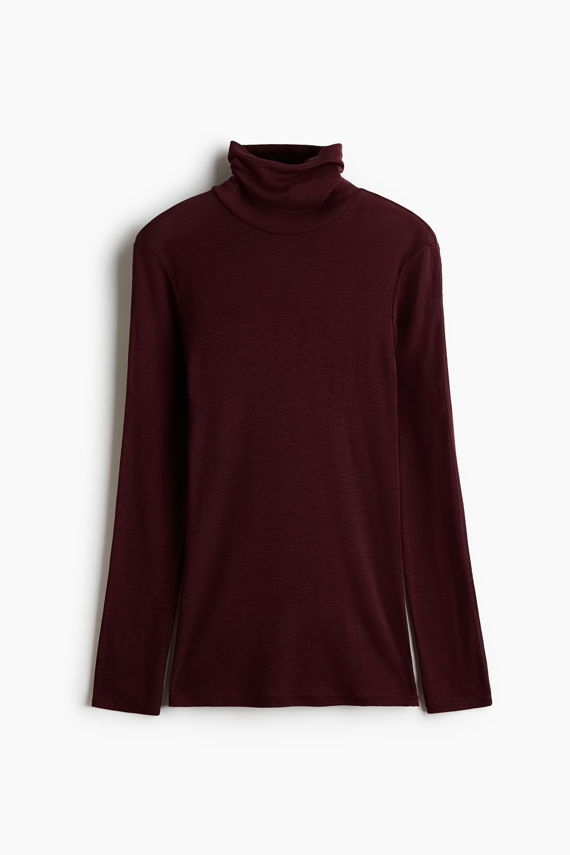 A fitted, dark burgundy turtleneck top in a fine rib knit is being worn by the model, who is crouching with arms crossed over one knee. The long-sleeved top is styled with wide-leg black trousers and black leather shoes.
