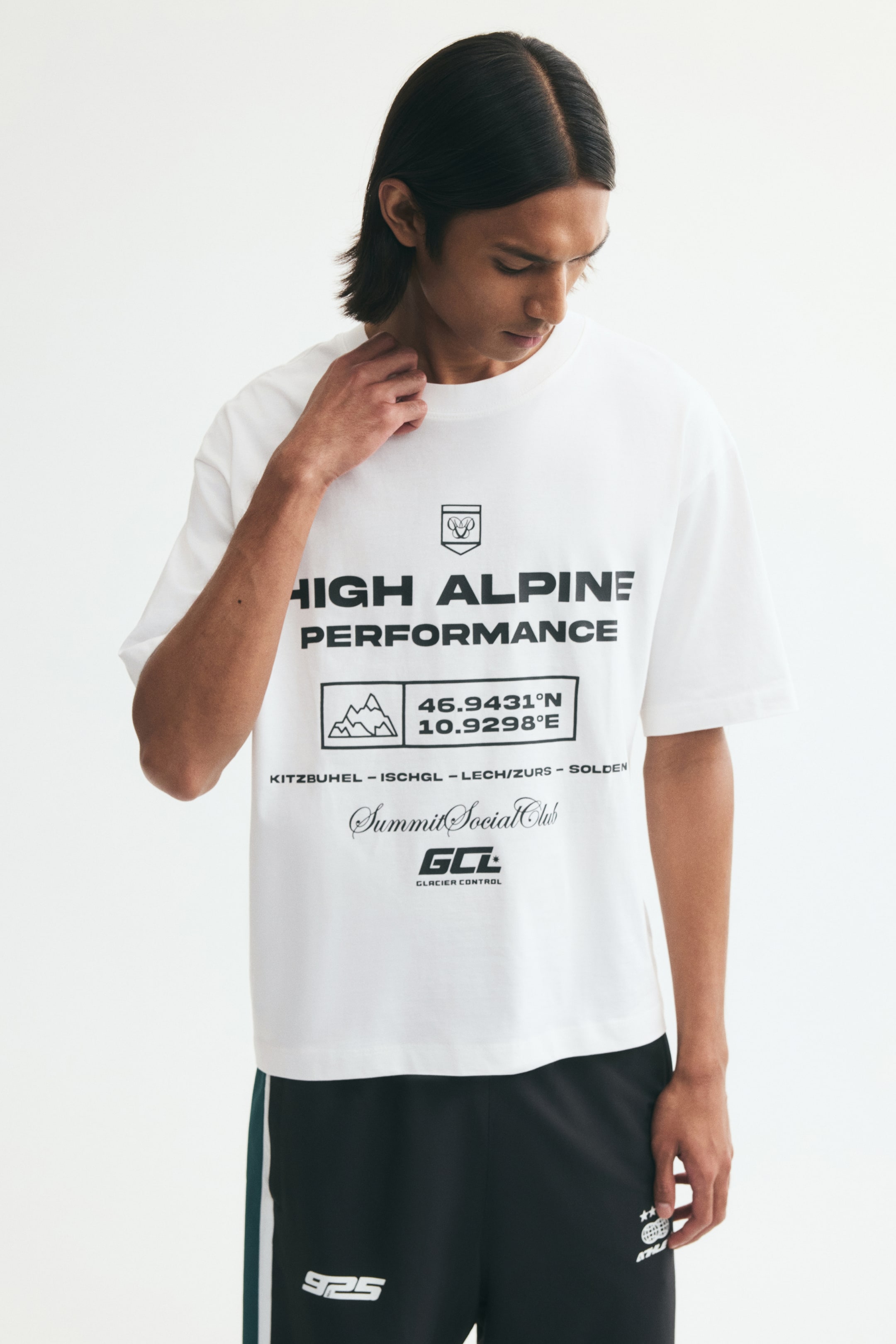 View larger image: A white, relaxed-fit T-shirt featuring a black graphic print on the front is worn by the model, who is touching the rib-trimmed neckline with one hand. The tee features dropped shoulders and a straight hem, and is paired with black athletic shorts.