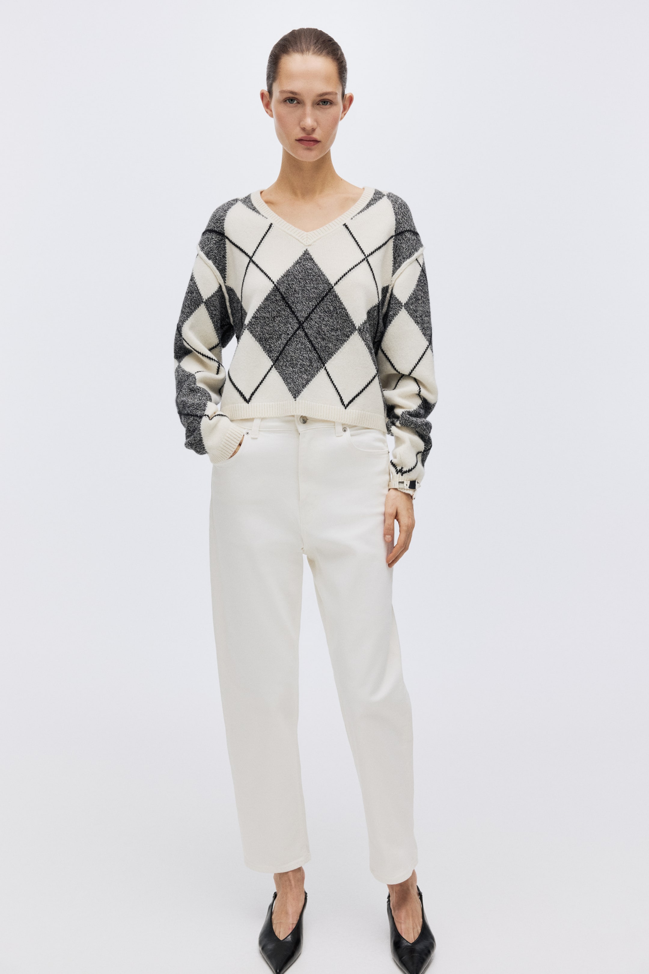 View larger image: A V-neck argyle sweater, featuring black and grey diamonds on an off-white base, is styled with white high-waisted pants. The sweater displays a cropped length and ribbed cuffs, while the pants offer a relaxed, cropped fit.