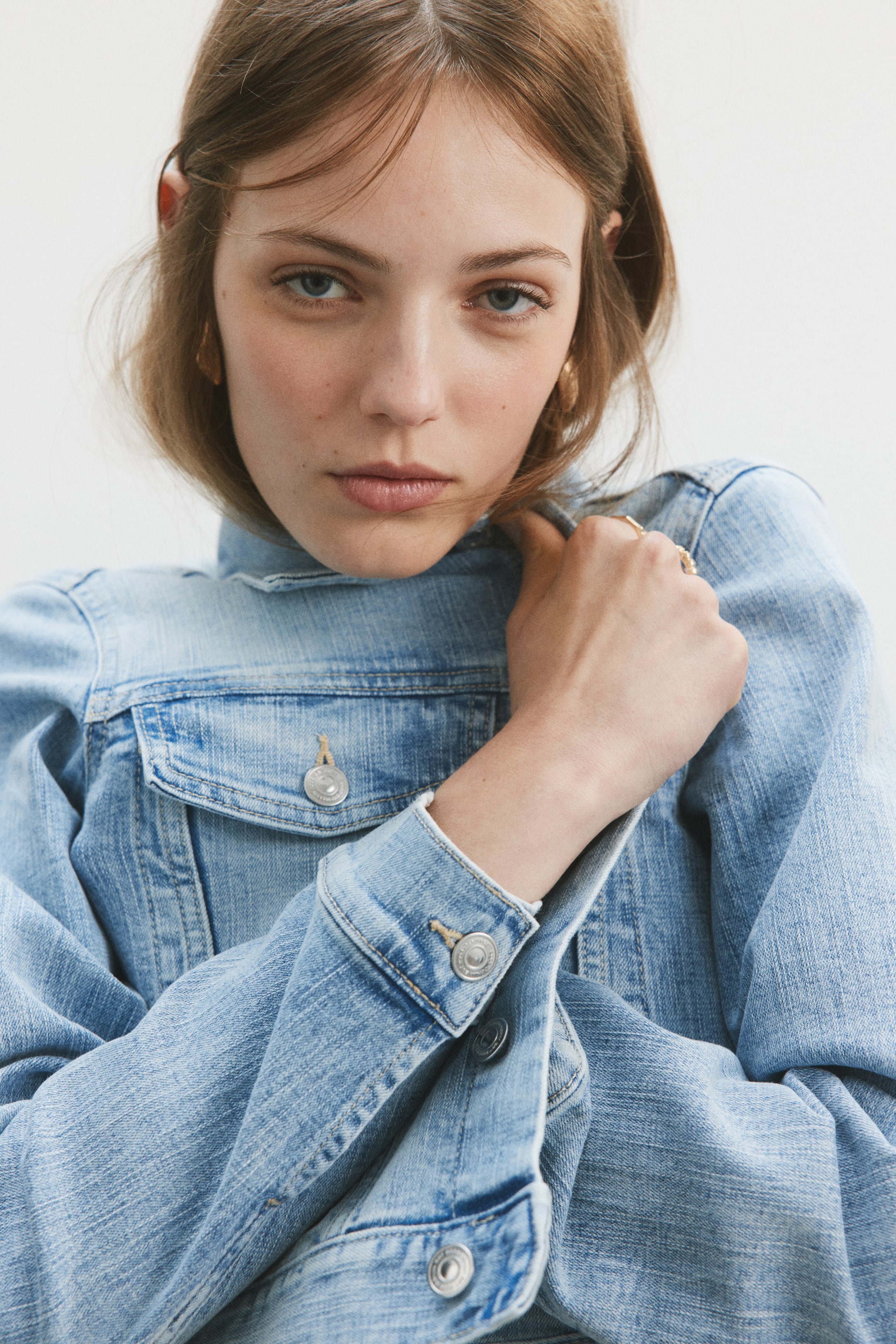View larger image: A tailored denim jacket, featuring a collar, buttons along the front, flap chest pockets with buttons, buttoned cuffs, and prominent seams, is worn by the model. The model's hand is adjusting the jacket's collar.
