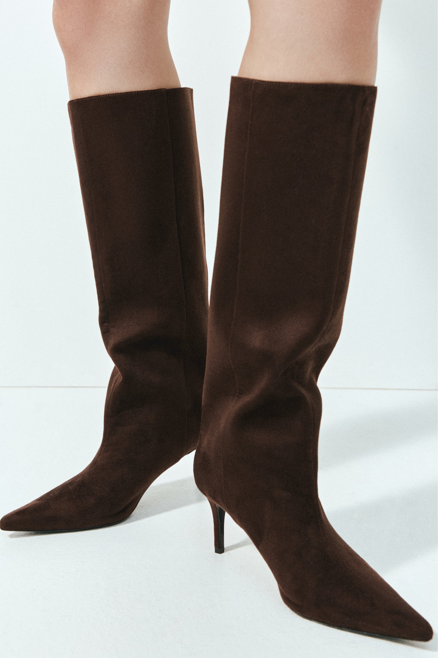 A dark brown boot is facing right, showcasing a pointed toe, a slender kitten heel, and a broad, knee-high shaft in a matte, suede-like finish. - 1