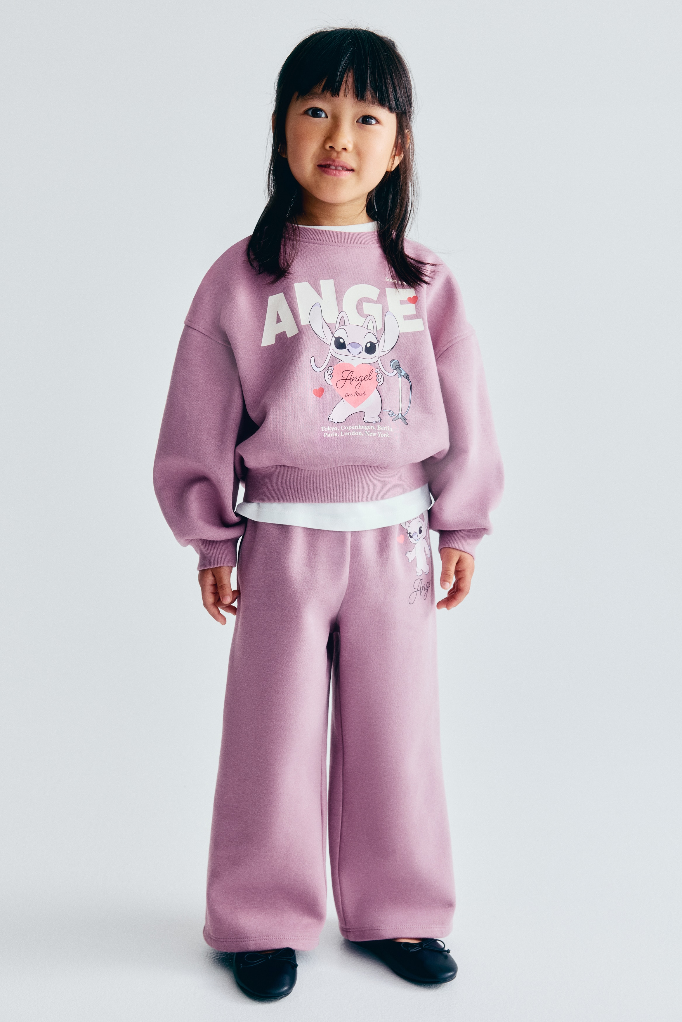 A two-piece purple loungewear set is shown from the front, featuring a cropped crew-neck sweatshirt and matching wide-leg trousers. The sweatshirt displays a large graphic of the character Angel beneath the word "ANGEL" printed in bold white letters, finished with ribbed cuffs and a ribbed hem. The trousers feature a smaller coordinating character graphic on the upper left thigh.