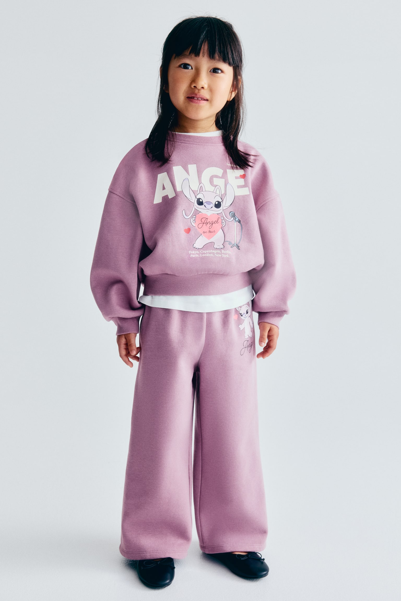 A two-piece purple loungewear set is shown from the front, featuring a cropped crew-neck sweatshirt and matching wide-leg trousers. The sweatshirt displays a large graphic of the character Angel below the word "ANGEL" printed in bold white letters, finished with ribbed cuffs and a ribbed hem. The trousers feature a smaller coordinating character graphic on the upper left thigh. - 1