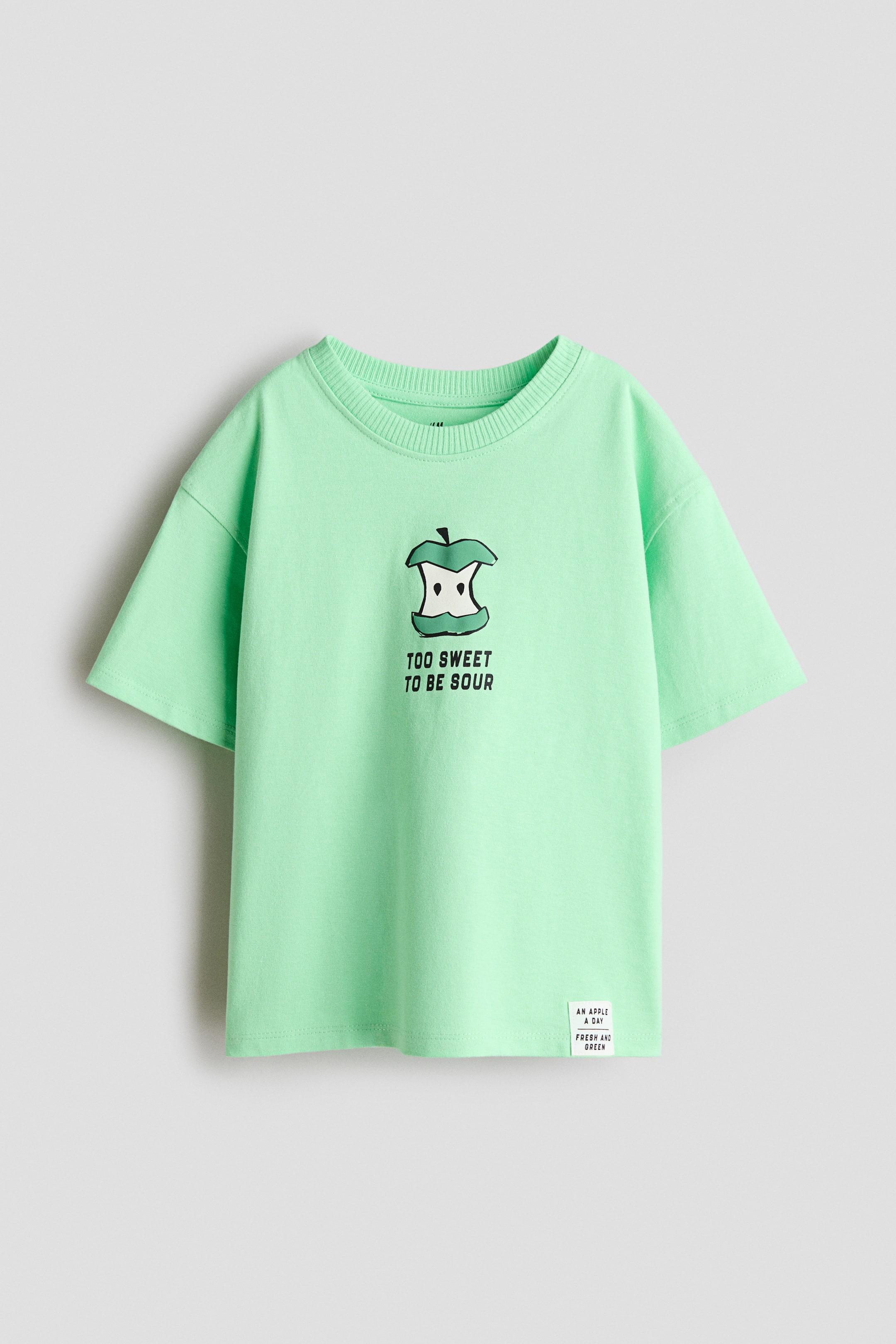 View larger image: A relaxed-fit T-shirt crafted from soft, light green cotton jersey is shown from the front, highlighting a round, ribbed neckline and dropped shoulders. The chest features a graphic print of a bitten apple core with the phrase "TOO SWEET TO BE SOUR", complemented by a small white label on the lower right front stating "AN APPLE A DAY FRESH AND GREEN".