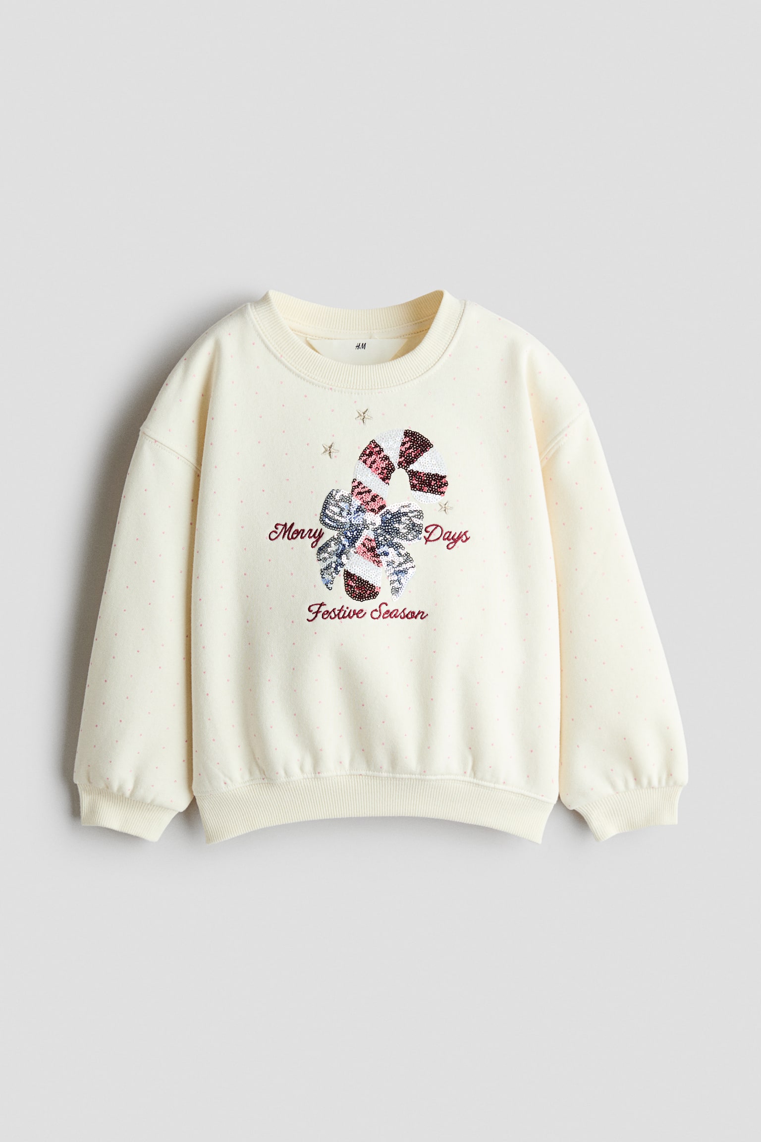 A cream-coloured sweatshirt is shown from the front, featuring a subtle pink polka dot pattern, dropped shoulders, and ribbed trim at the crew neck, cuffs, and hem. The front features a sequined candy cane graphic adorned with a sequined bow, surrounded by embroidered gold stars and red script reading "Merry Days Festive Season."