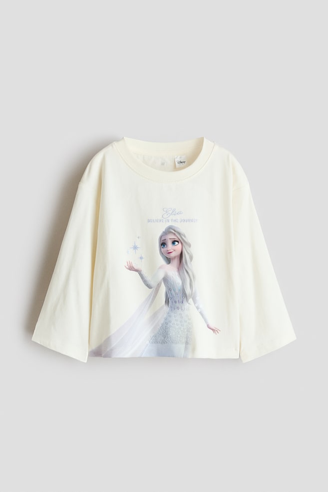 En cremefarvet langÃ¦rmet T-shirt er vist forfra og fremhÃ¦ver en rund halsudskÃ¦ring med smal ribkant og brede, afslappede Ã¦rmer. Forsiden viser et trykt motiv af karakteren Elsa, med teksten "Elsa" og "BELIEVE IN THE JOURNEY" trykt over illustrationen. - 1