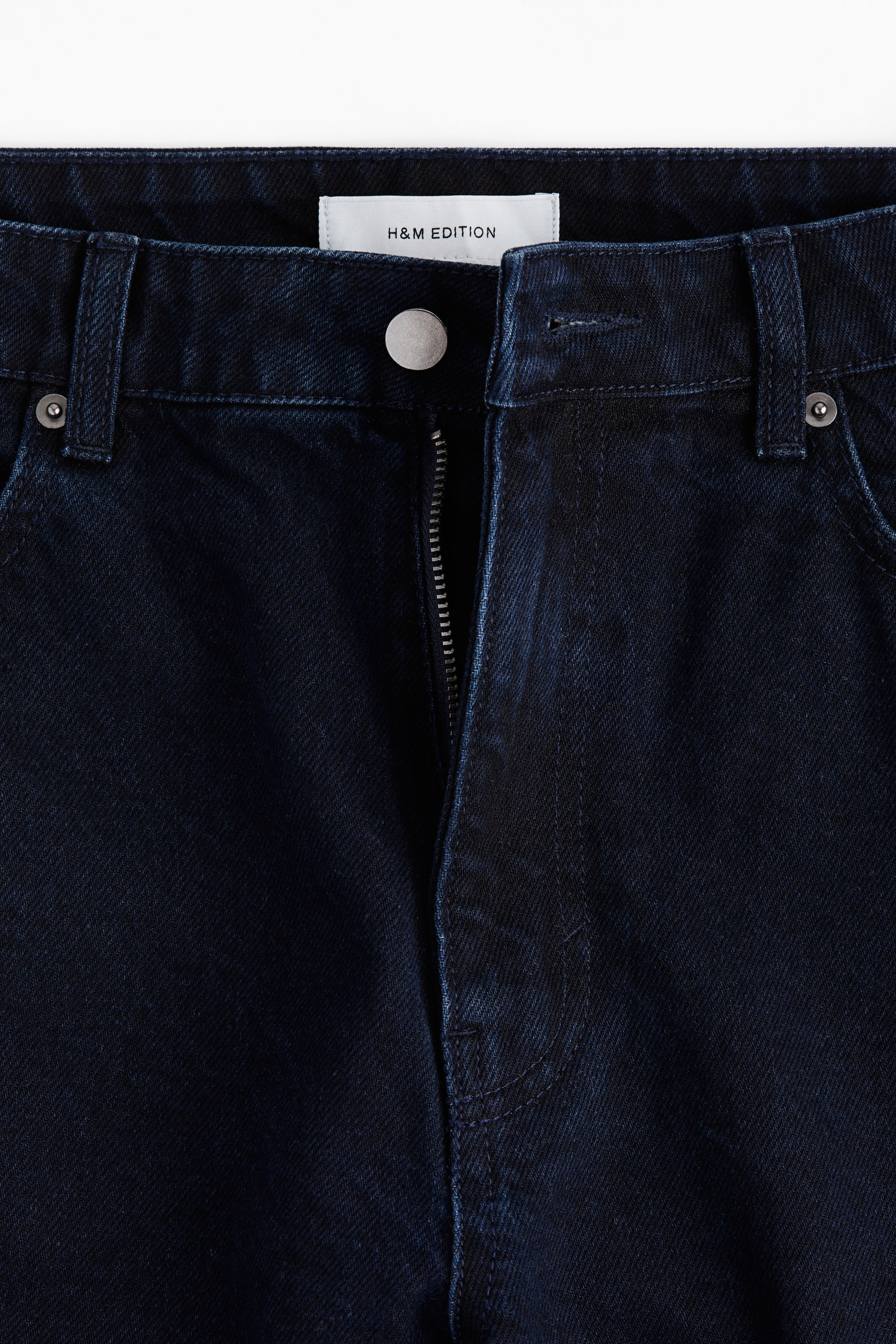 Voir une image plus grande: Ce jean à 5 poches en denim de coton rigide bleu foncé est présenté de face, avec une taille régulière et une braguette partiellement dézippée avec une fermeture à bouton. Un clou en métal en forme de dôme est visible sur la hanche gauche, faisant partie des clous détaillant les côtés extérieurs des jambes droites.