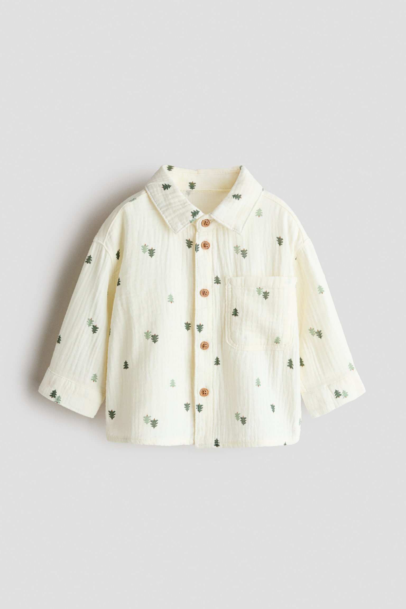 A cream-coloured, long-sleeved button-up shirt in textured crinkle fabric featuring a small, scattered green pine tree pattern. Shown from the front, the shirt showcases a classic collar, a single chest patch pocket, and five brown buttons down the centre placket.