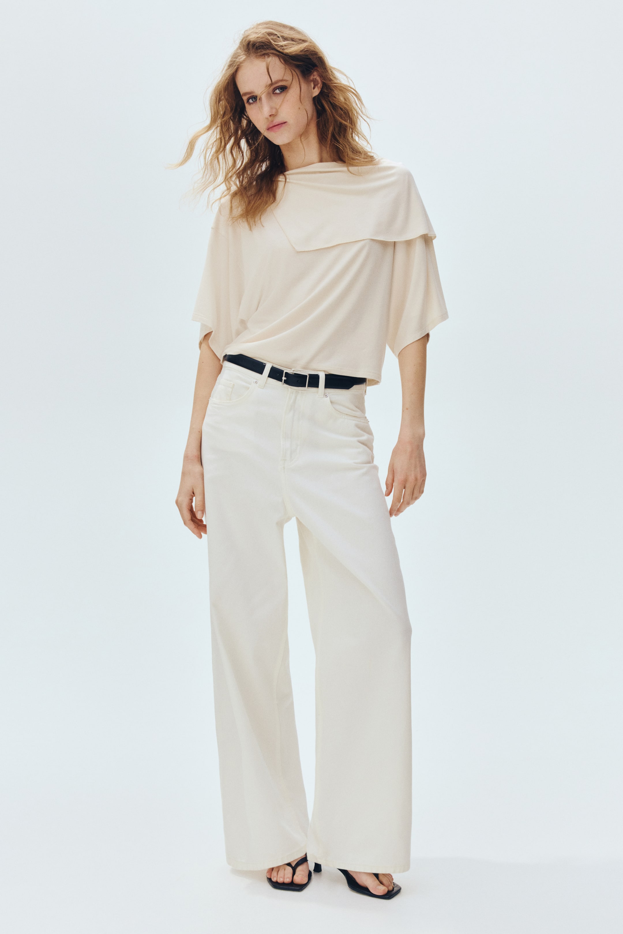 Ingrandisci l'immagine: Un top beige chiaro a vestibilità ampia presenta un collo a cascata drappeggiato e maniche corte larghe. Viene abbinato a pantaloni a vita alta, a gamba larga di colore bianco sporco, stretti da una cintura nera.