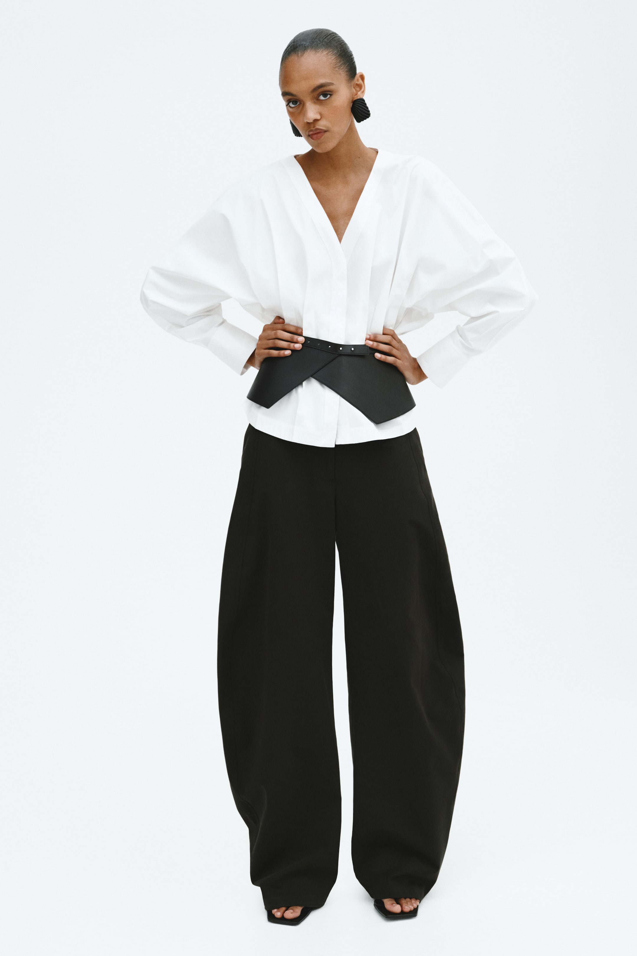 Grotere afbeelding bekijken: Een witte oversized blouse, gestyled met een V-hals en brede manchetten, wordt in de taille aangesnoerd door een zwarte brede asymmetrische riem met studs. Zwarte wijde broek maakt het ensemble compleet en wordt gecombineerd met zwarte open teen platte sandalen.