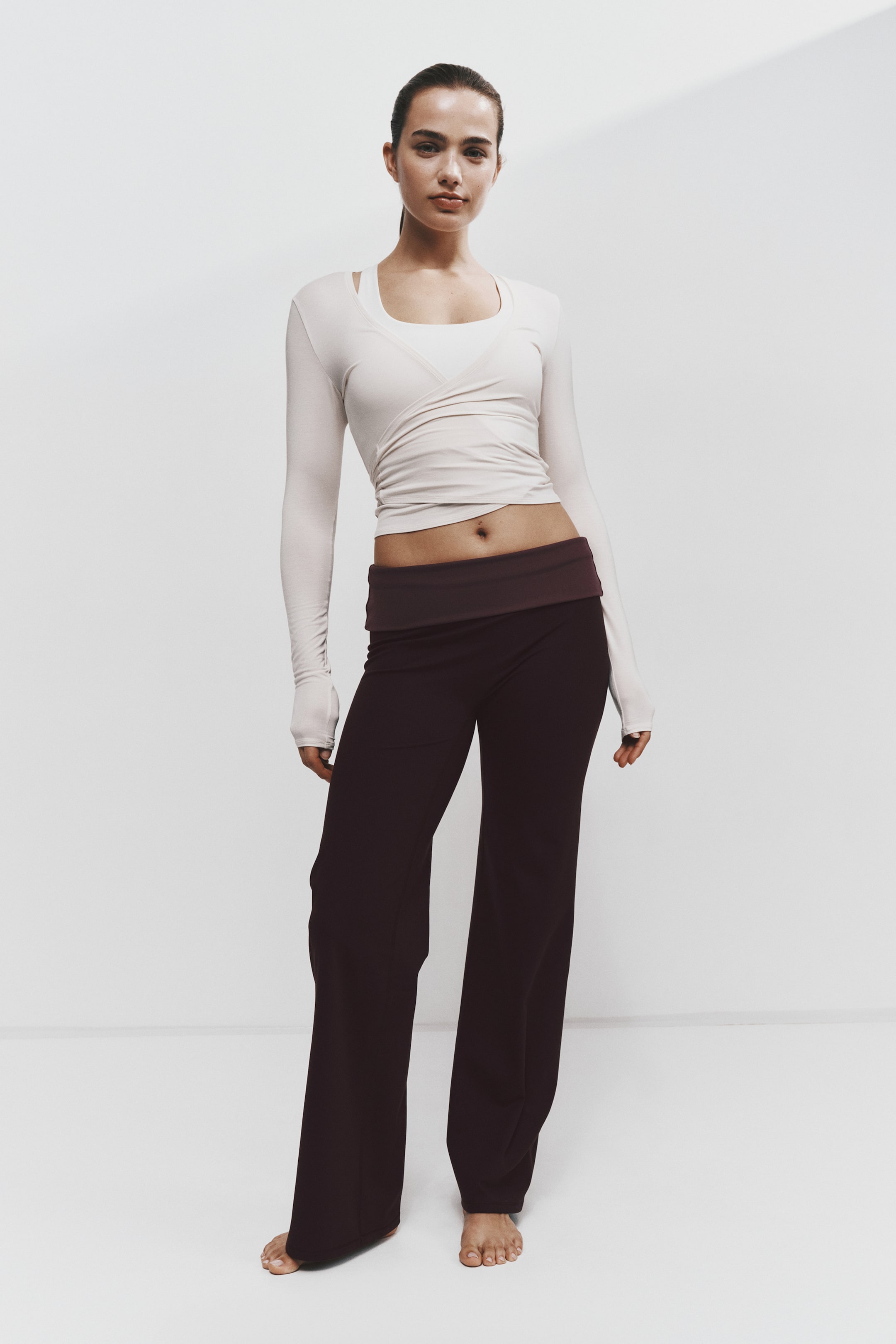 Se større billede: En model bærer en lys beige wrap-top med lange ærmer og tommelfingerhuller, lagt over en matchende tanktop, kombineret med mørke bordeauxfarvede wide-leg-bukser med fold-over linning. Sættet vises forfra og fremhæver toppens cropped, draperede design og buksernes afslappede silhuet.