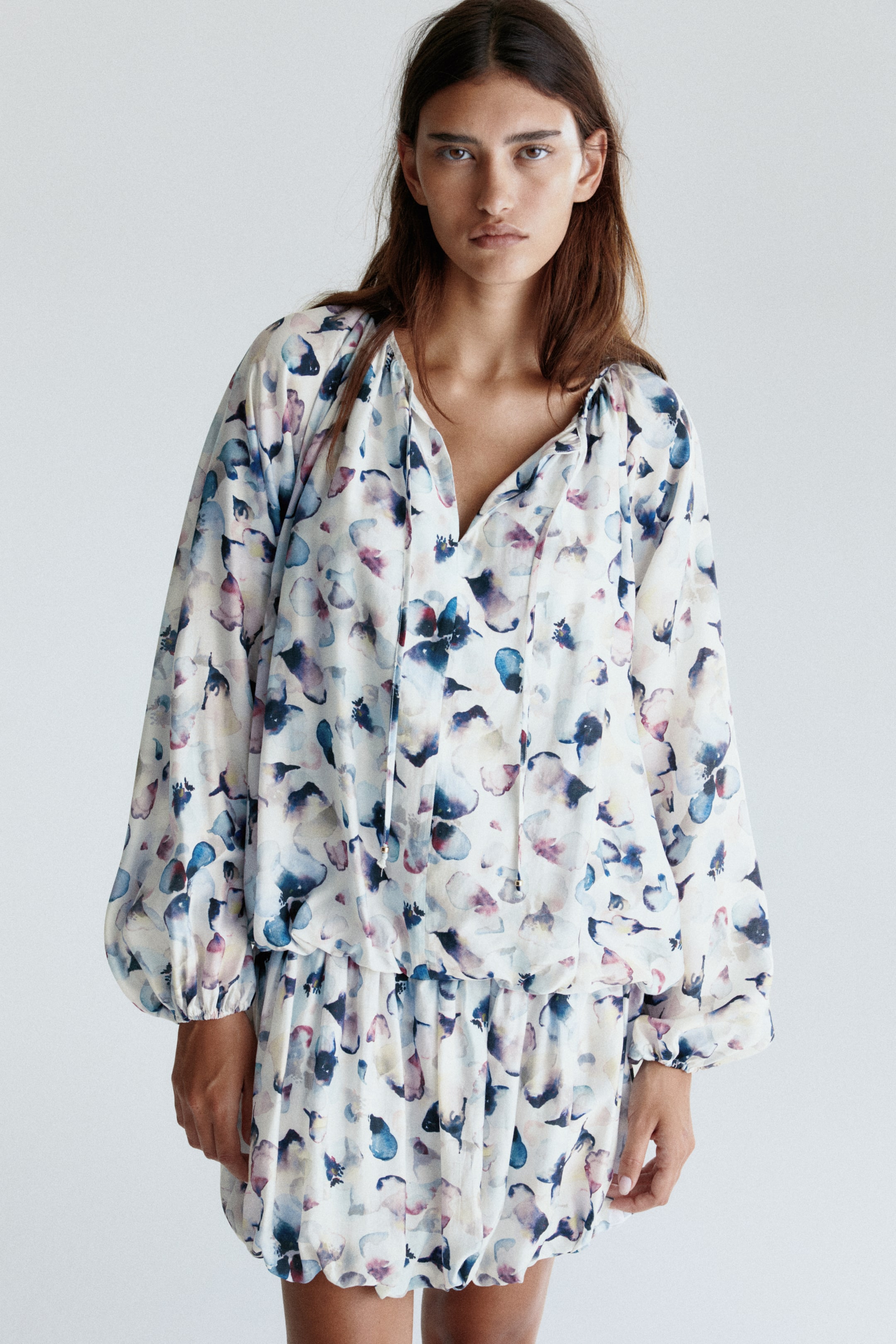 View larger image: A white dress is featuring an all-over watercolor-style abstract floral print in shades of blue and purple. It is designed with a V-neckline and tie details, long voluminous sleeves with elasticated cuffs, and a gathered elasticated waist creating a blouson top and a matching mini-skirt with an elasticated hem.