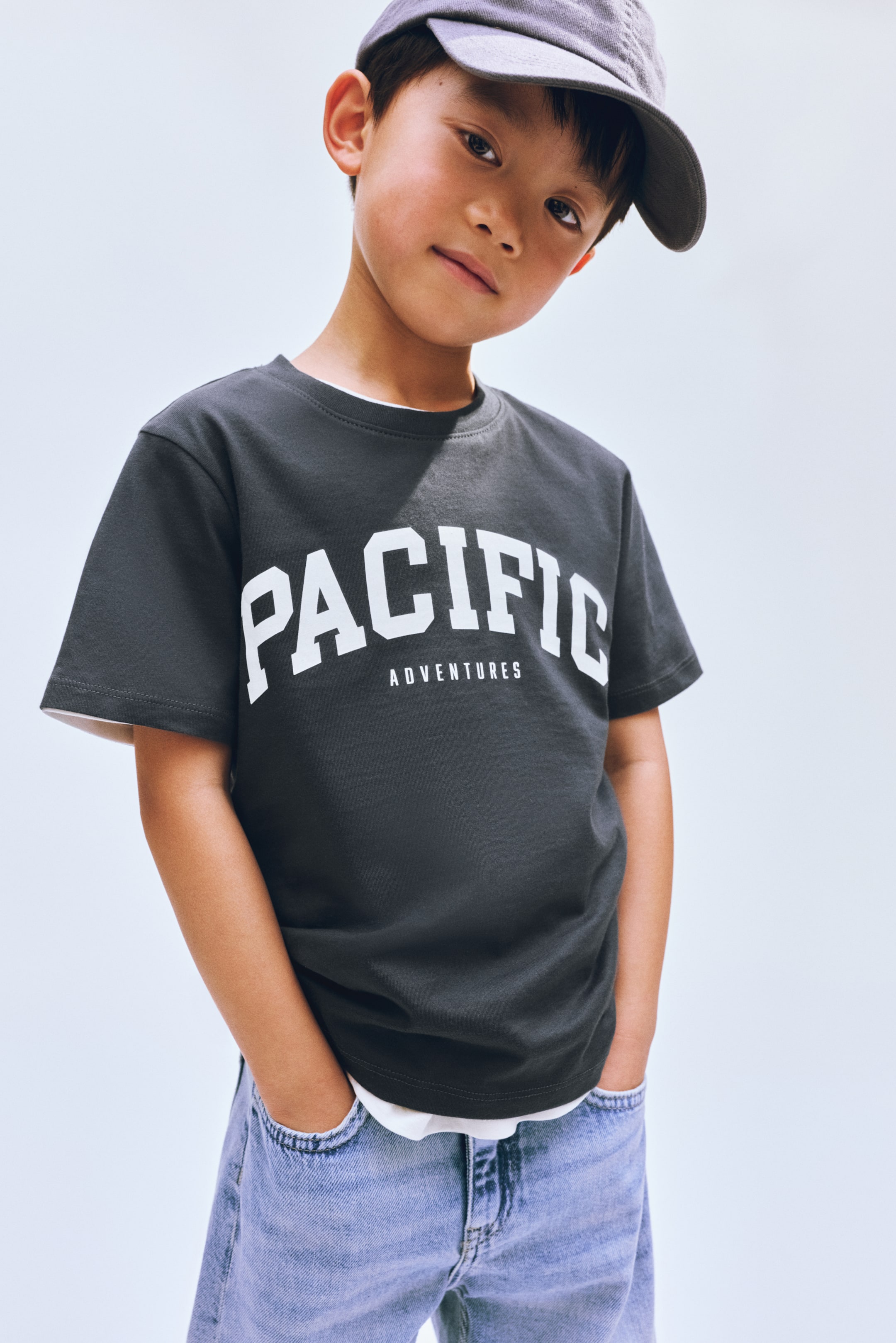 View larger image: A dark grey short-sleeved t-shirt showcases a white "PACIFIC ADVENTURES" graphic on the front, with "PACIFIC" prominently arched above "ADVENTURES." It is layered over a white t-shirt, visible at the crew neck and hem.