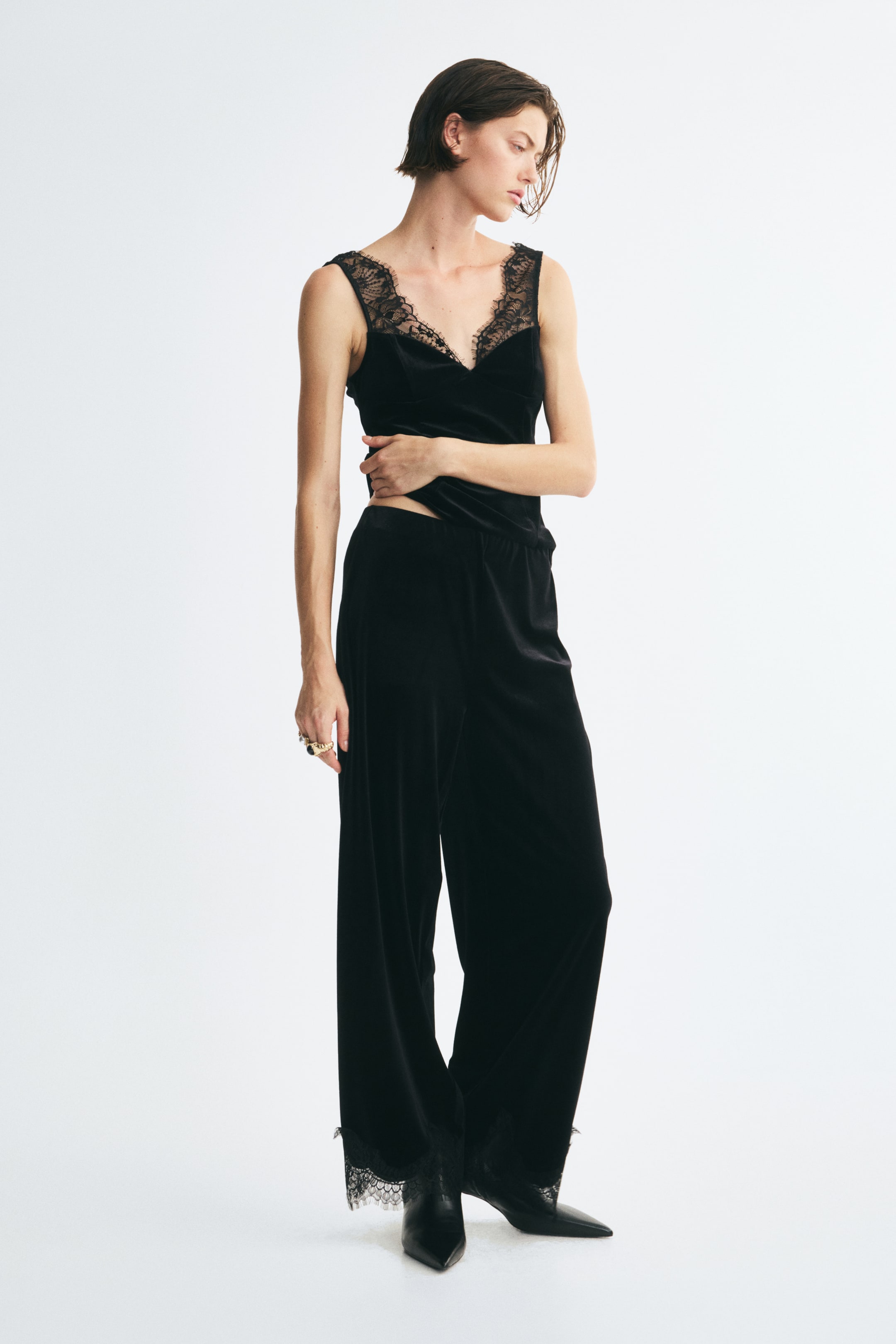 Grotere afbeelding bekijken: Zwarte velours broek met een losse pasvorm, rechte pijpen en geschulpte kanten afwerking aan de zoom wordt gedragen door het model. De broek is gestyled met een bijpassende zwarte camisole met kanten afwerking en zwarte laarzen met puntneus.