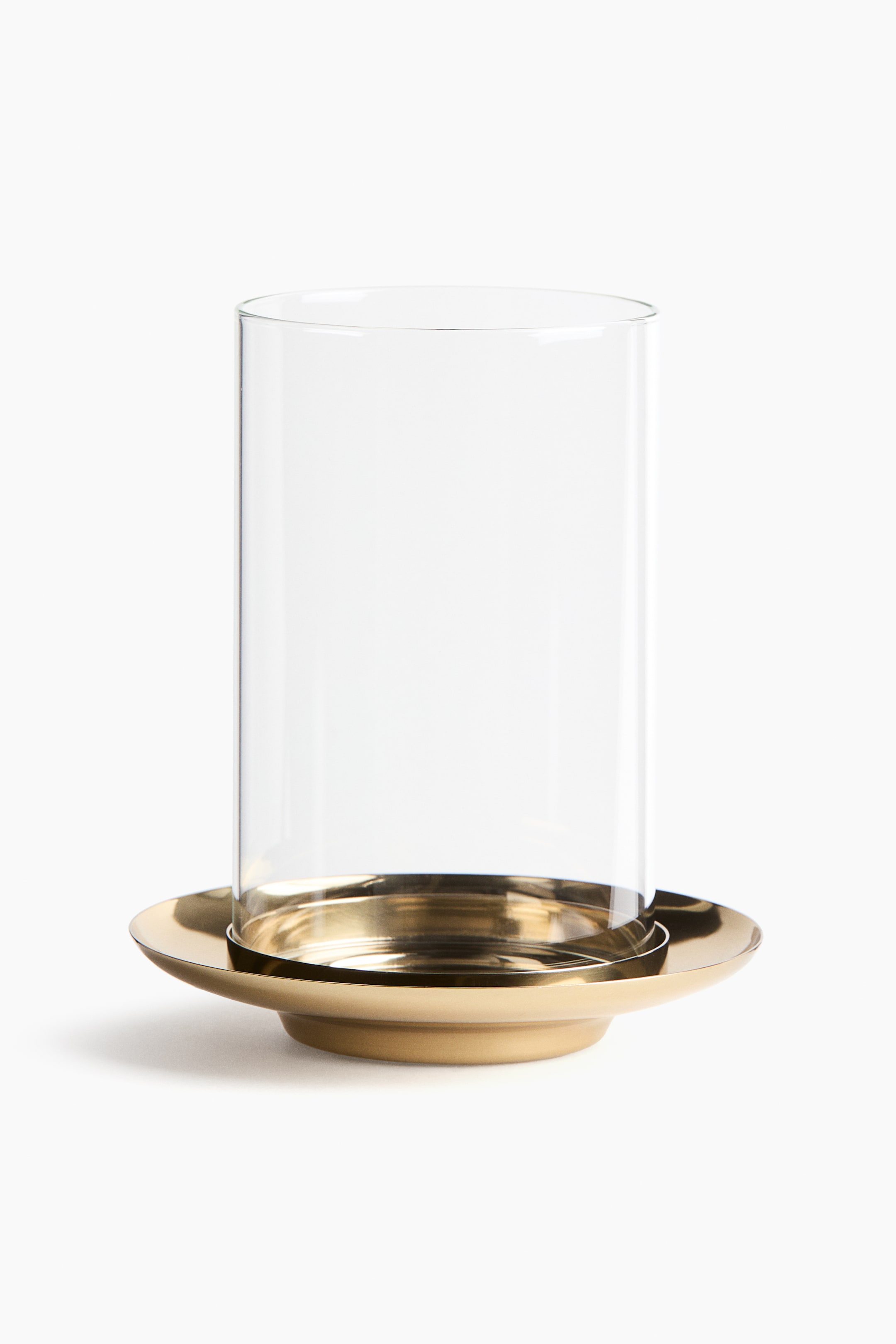 View larger image: A candle holder features a tall, clear glass cylinder positioned centrally within a round brass base. The brass base has a raised inner section and a wider, flat outer rim.